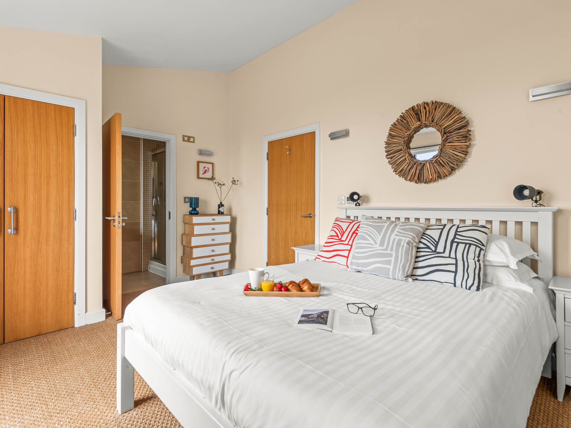 Bright bedroom with white bed, patterned cushions and driftwood-framed mirror, with open door to en-suite shower