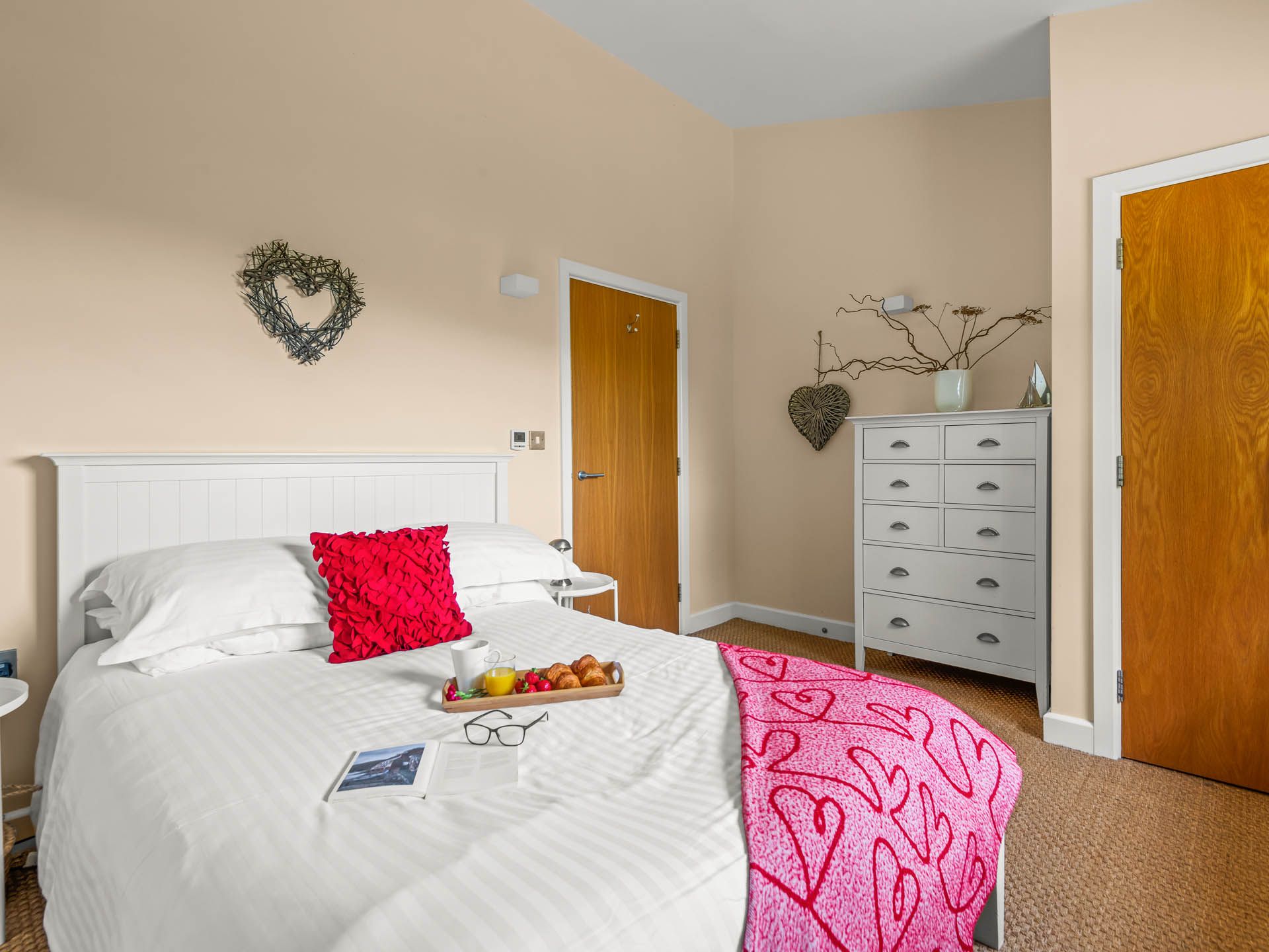 Bright bedroom with white bed, red cushion and pink throw, wooden doors and grey chest of drawers