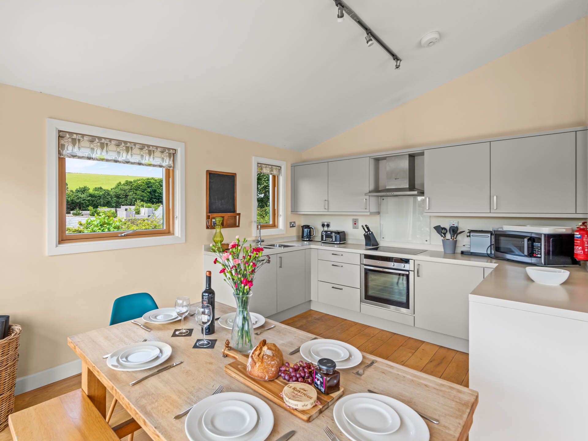 Modern open-plan kitchen and dining area with grey cabinets, wooden table set for a meal, and a countryside view through the window