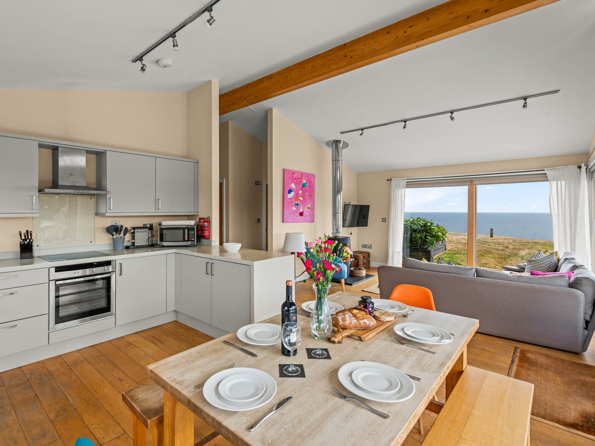 Open-plan kitchen and dining area with grey cabinets and a set table, leading to a lounge with large sliding doors overlooking the sea