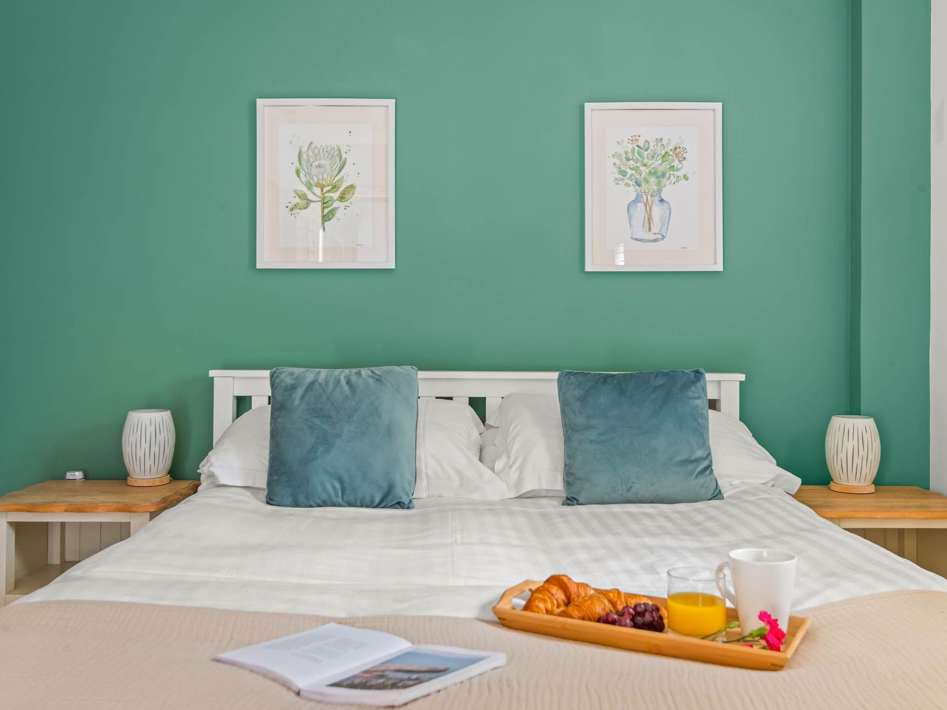 Neatly made bed with teal cushions against a turquoise wall, framed botanical prints above and breakfast tray on the duvet