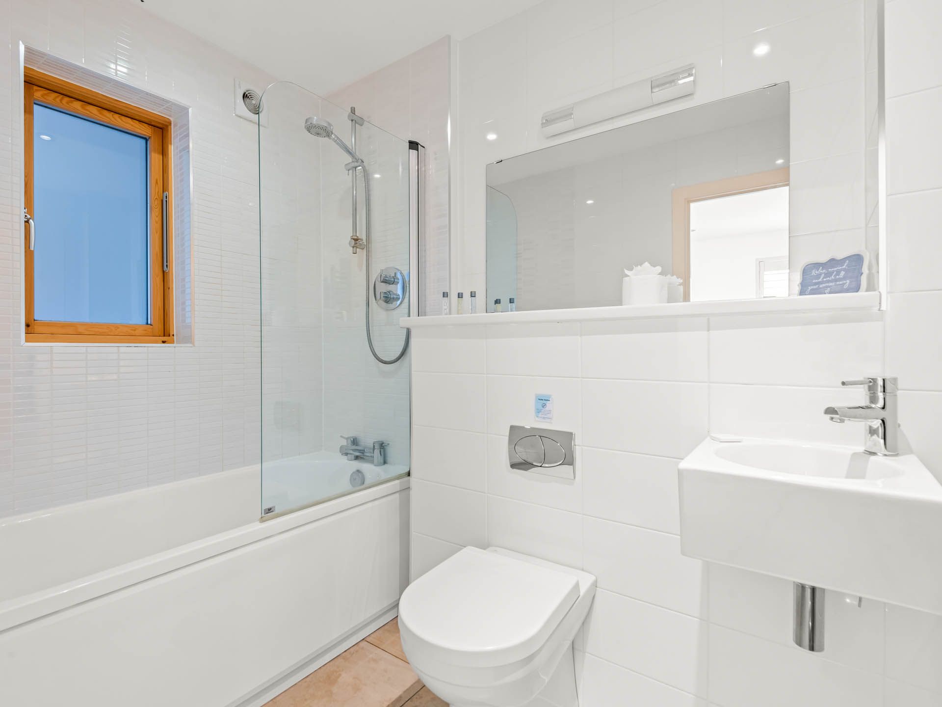 Bright white tiled bathroom with shower over bath, wall-hung toilet, vanity sink and wooden-framed window