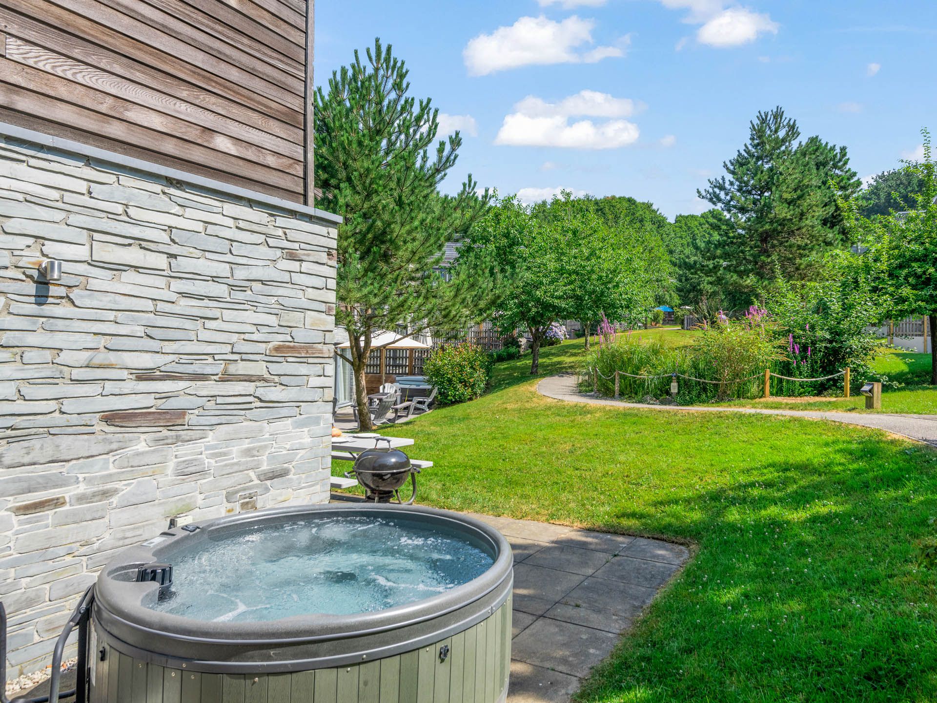 Outdoor hot tub on a paved patio beside a stone-and-wood clad building, overlooking a green garden with a winding path and trees