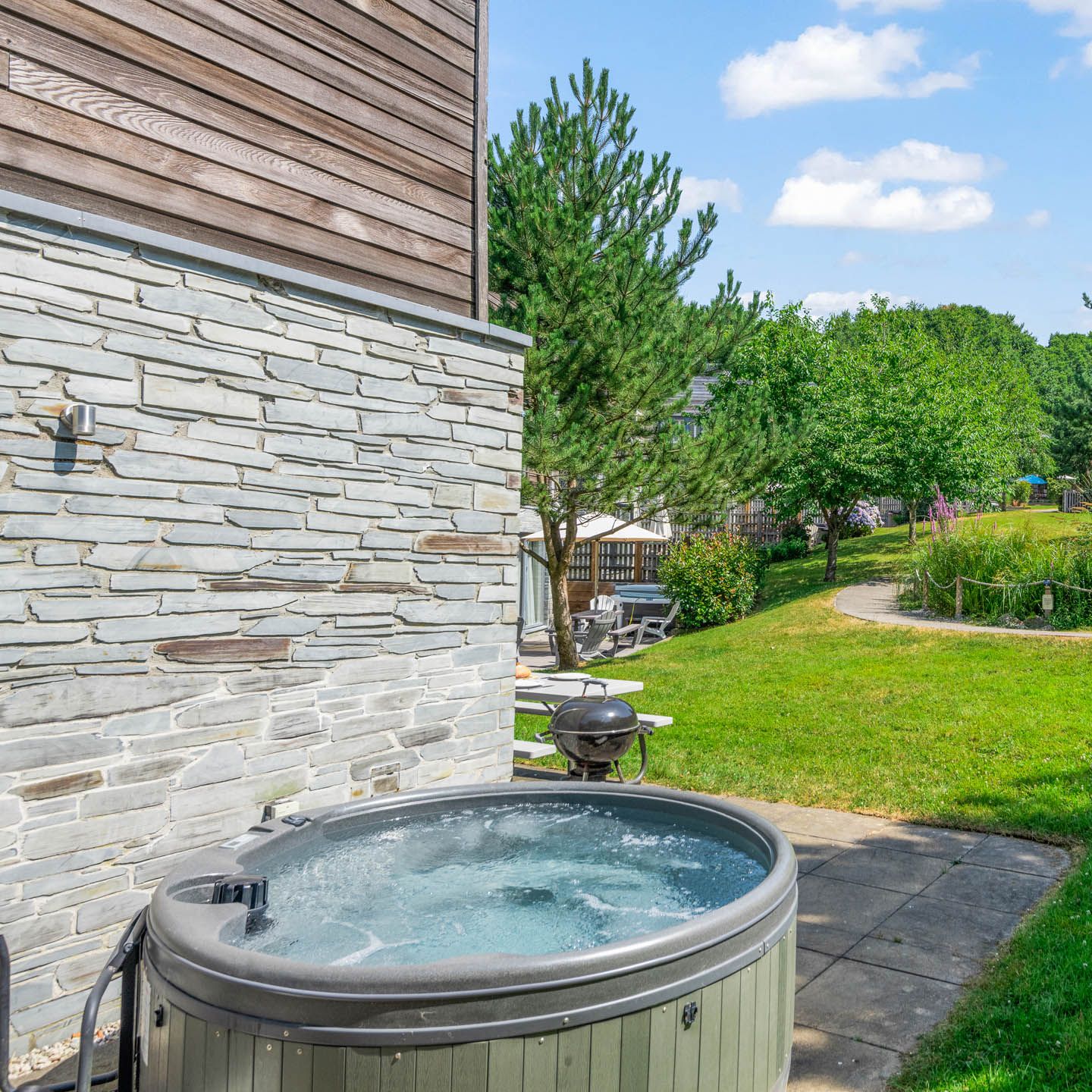 Outdoor hot tub on a paved patio beside a stone-and-wood clad building, overlooking a green garden with a winding path and trees