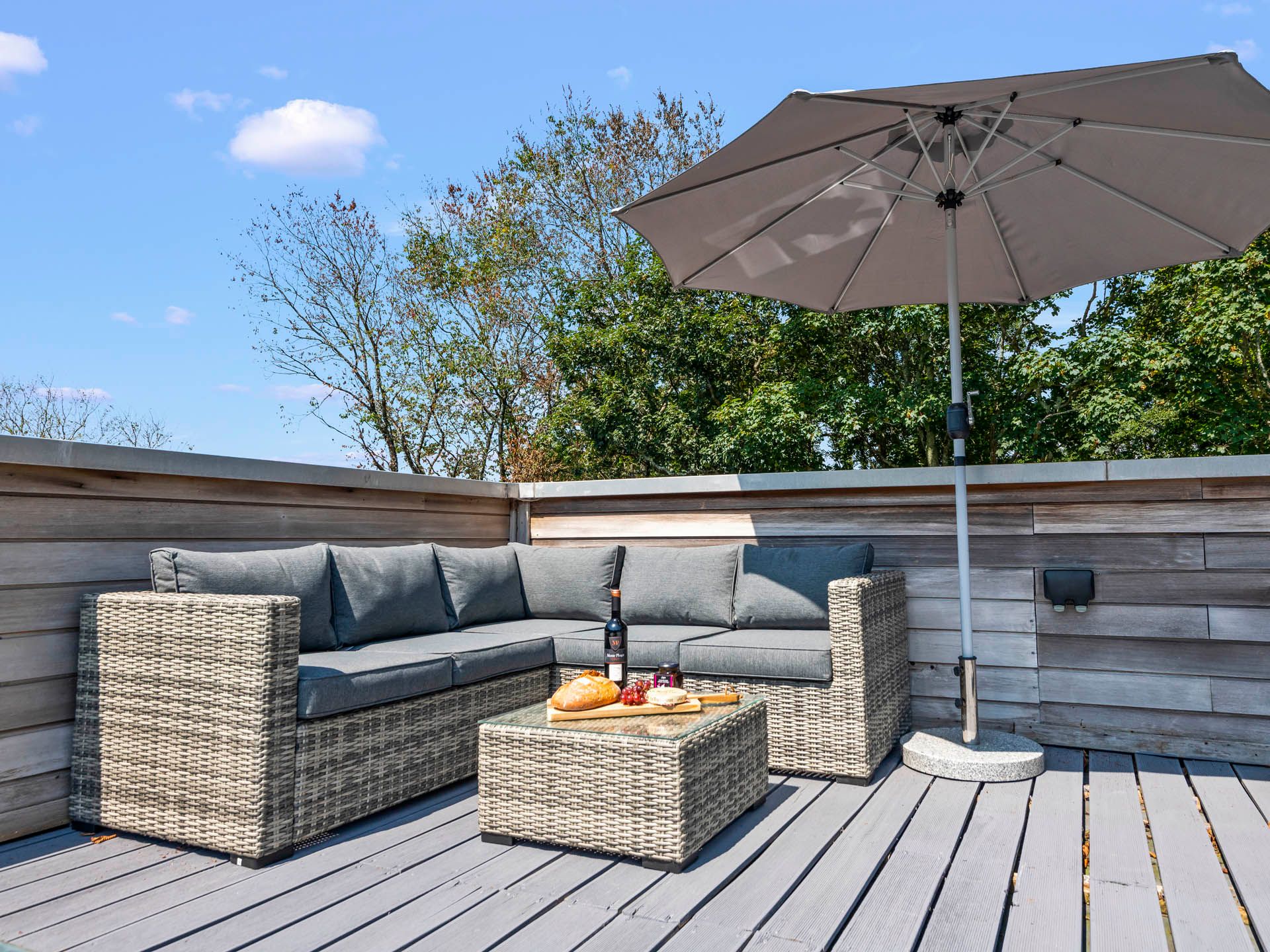 Outdoor patio seating area with wicker corner sofa, grey cushions and large parasol on a wooden deck