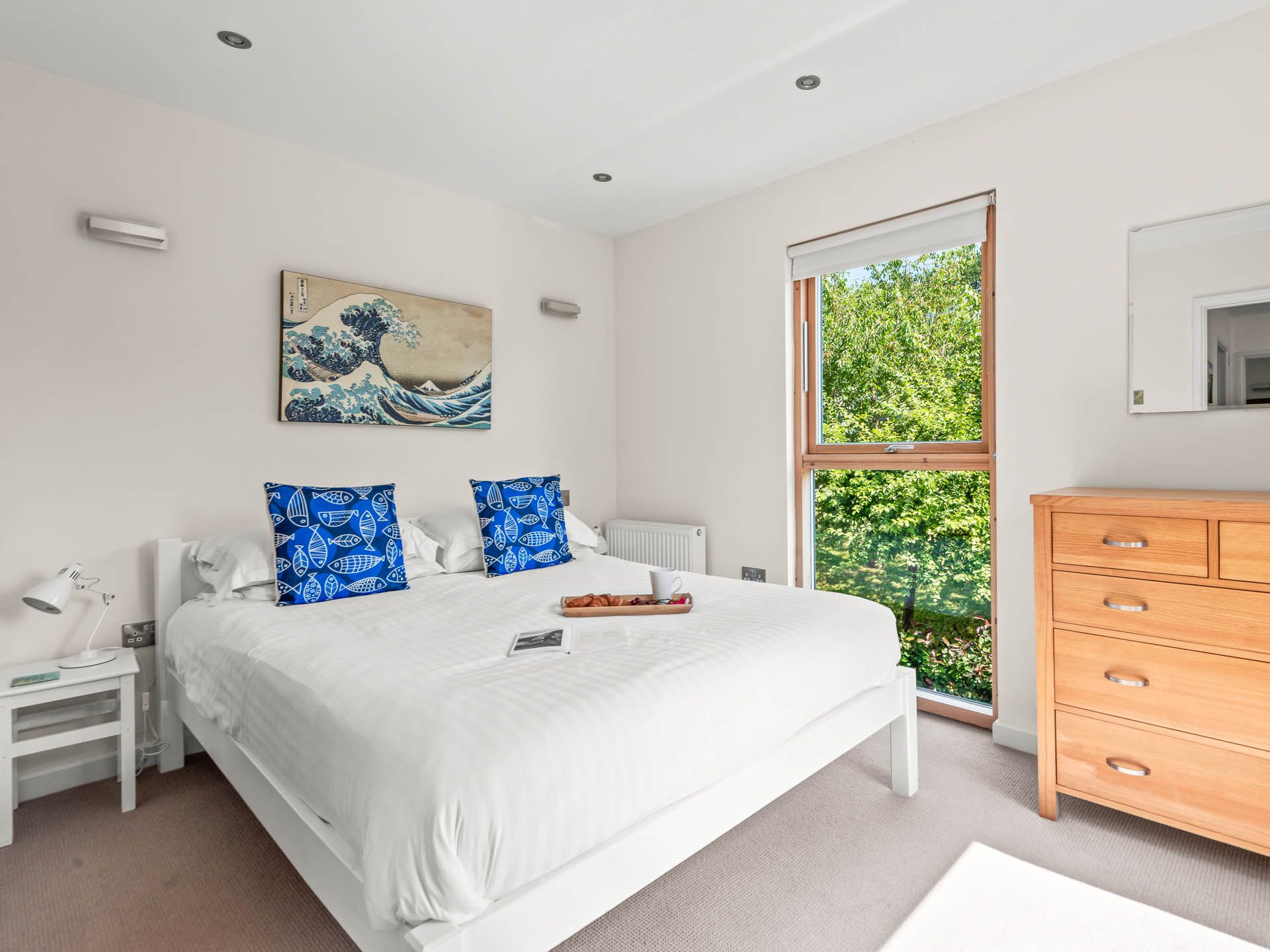 Bright double bedroom with white bedding, blue patterned cushions, wooden chest of drawers and a large window overlooking green trees
