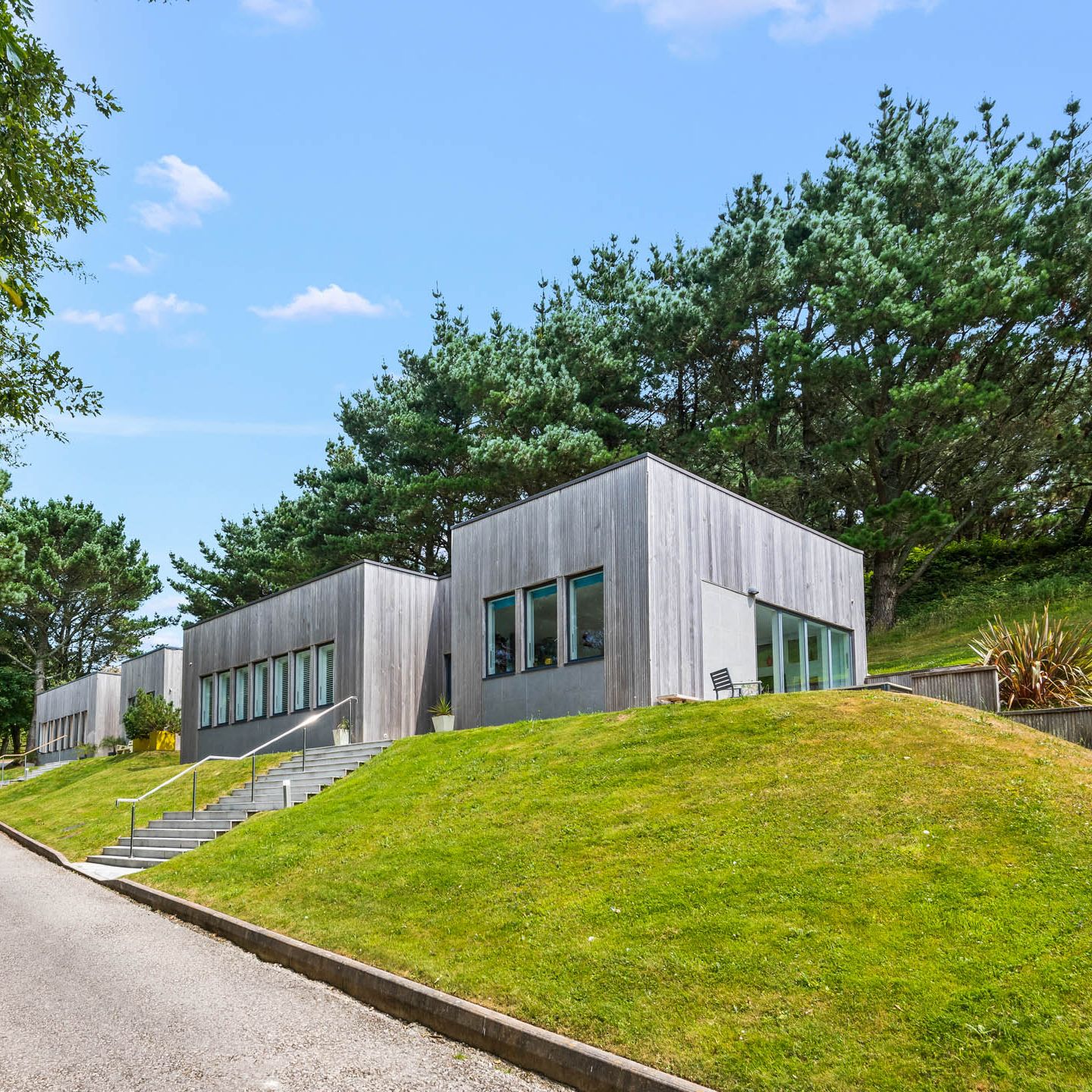 Modern timber-clad building on a grassy hillside with a path and surrounding trees under a blue sky