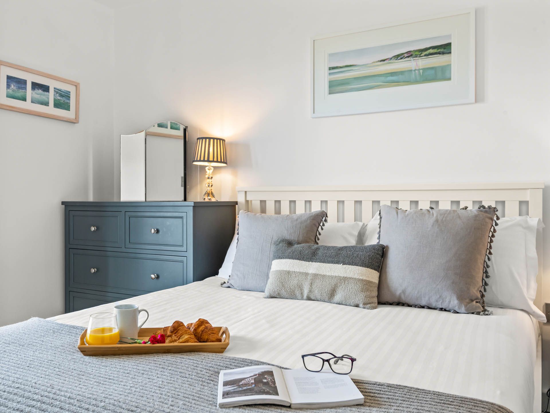 Cosy bedroom with white bed, grey cushions, breakfast tray and bedside lamp next to a blue chest of drawers
