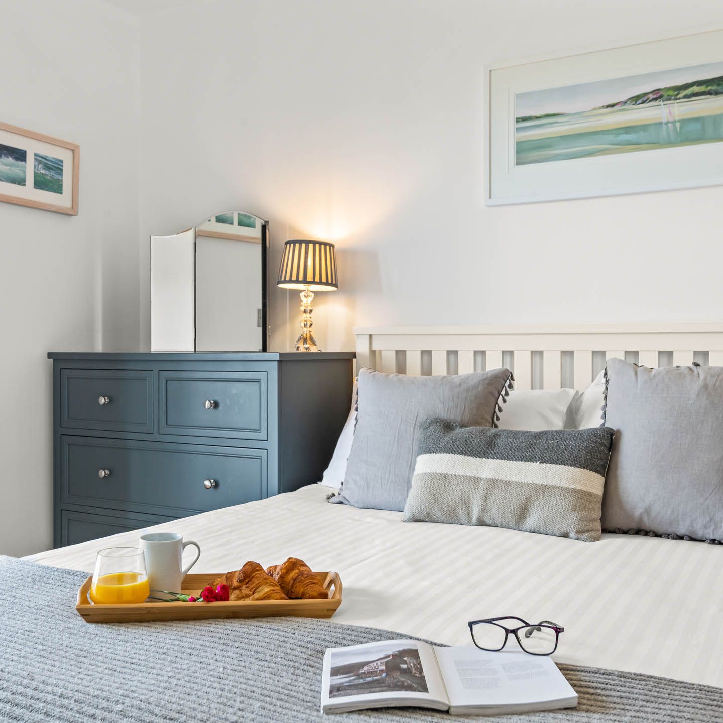 Cosy bedroom with white bed, grey cushions, breakfast tray and bedside lamp next to a blue chest of drawers