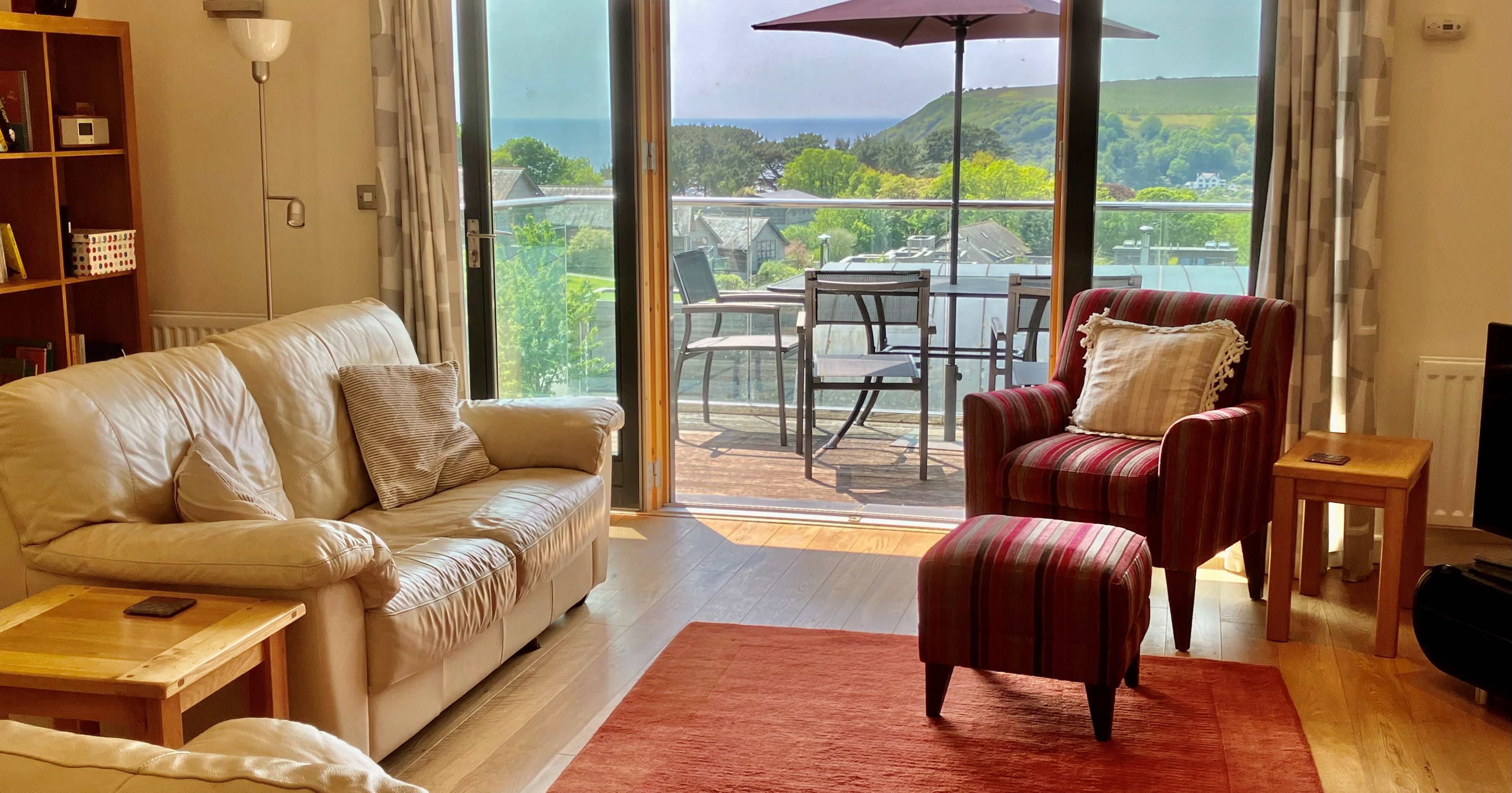 Bright living room with leather sofa, striped armchair, coffee tables and sliding glass doors opening to a patio with outdoor seating and umbrella, overlooking a green hillside.