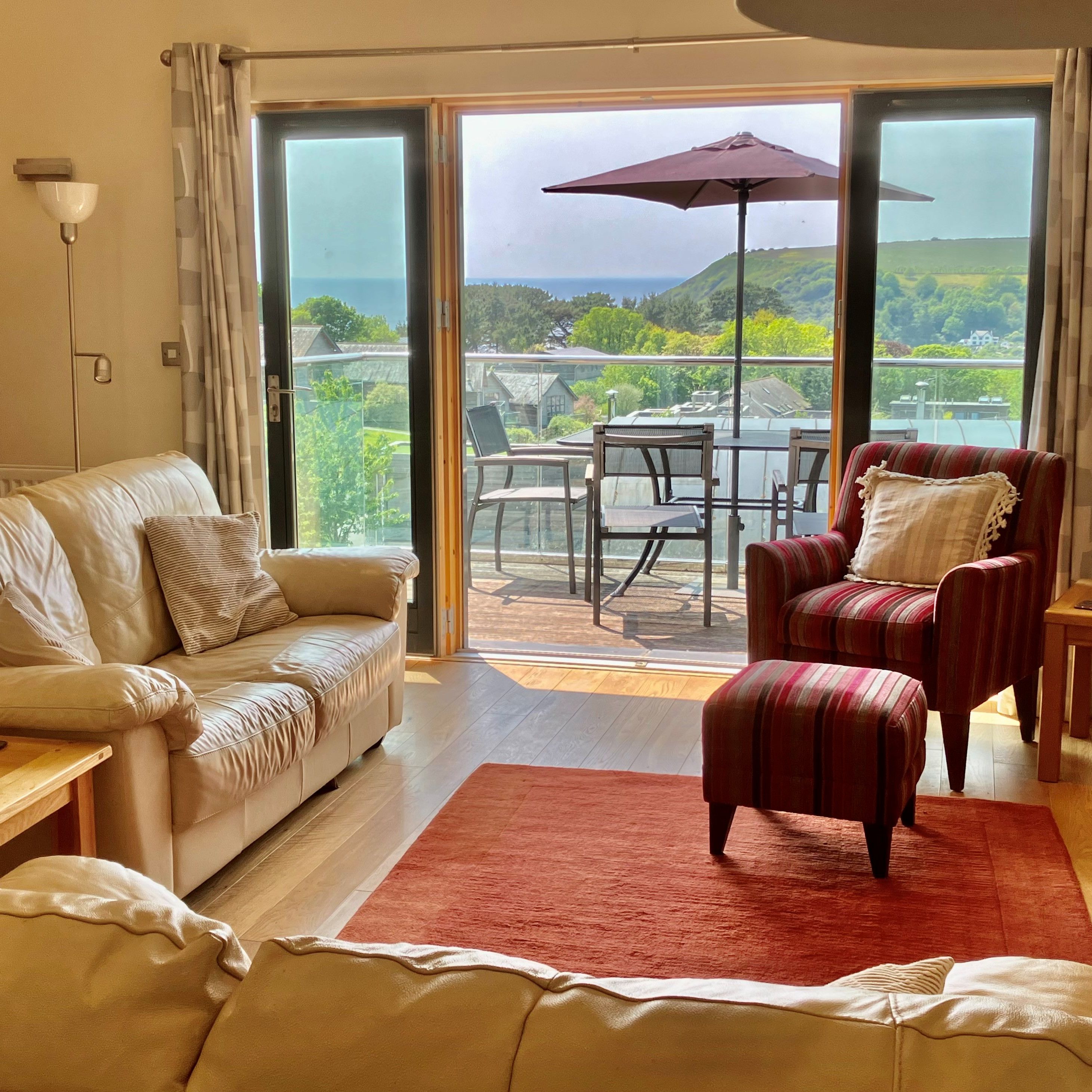 Bright living room with leather sofa, striped armchair, coffee tables and sliding glass doors opening to a patio with outdoor seating and umbrella, overlooking a green hillside.