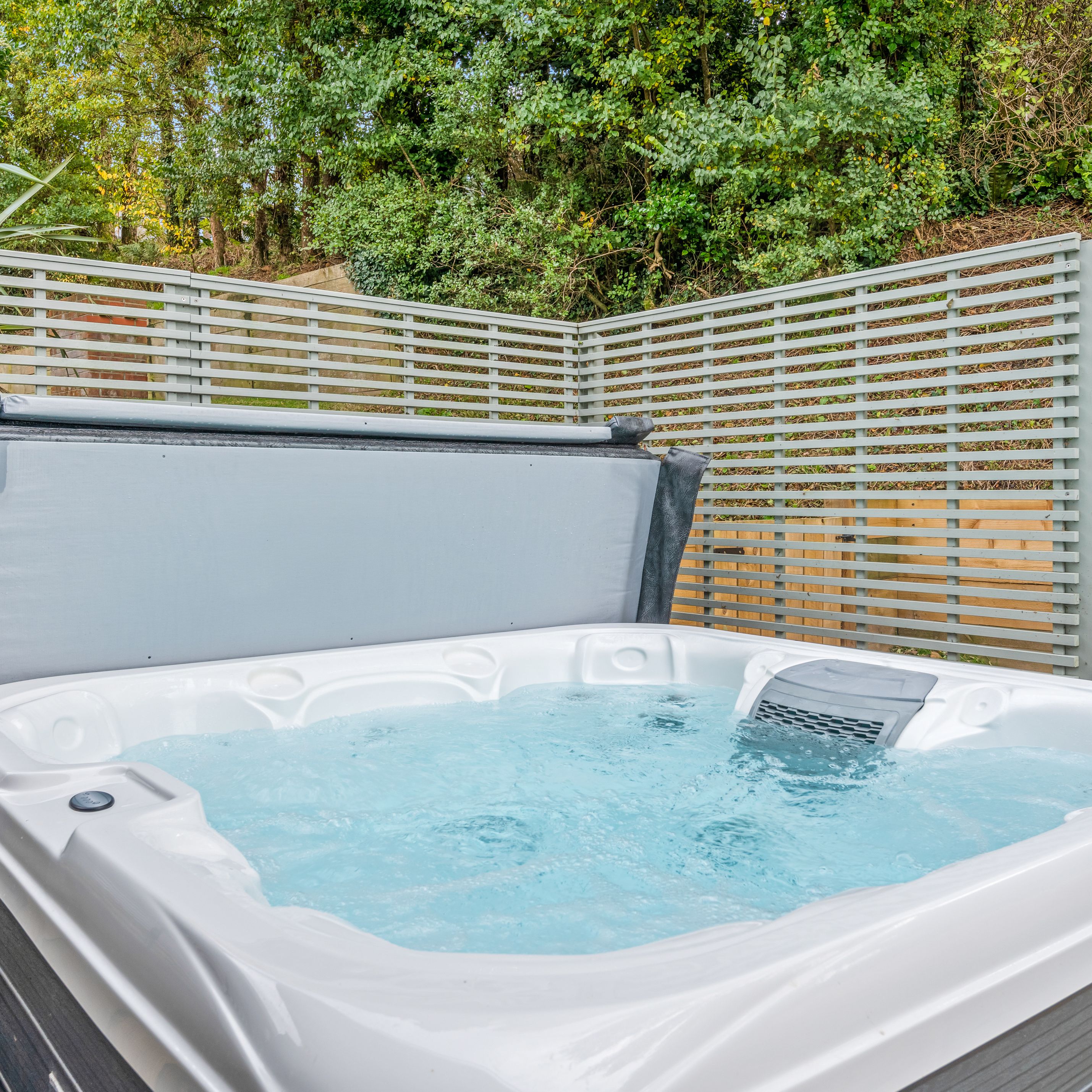Outdoor hot tub with bubbling water beside a grey slatted privacy screen and trees