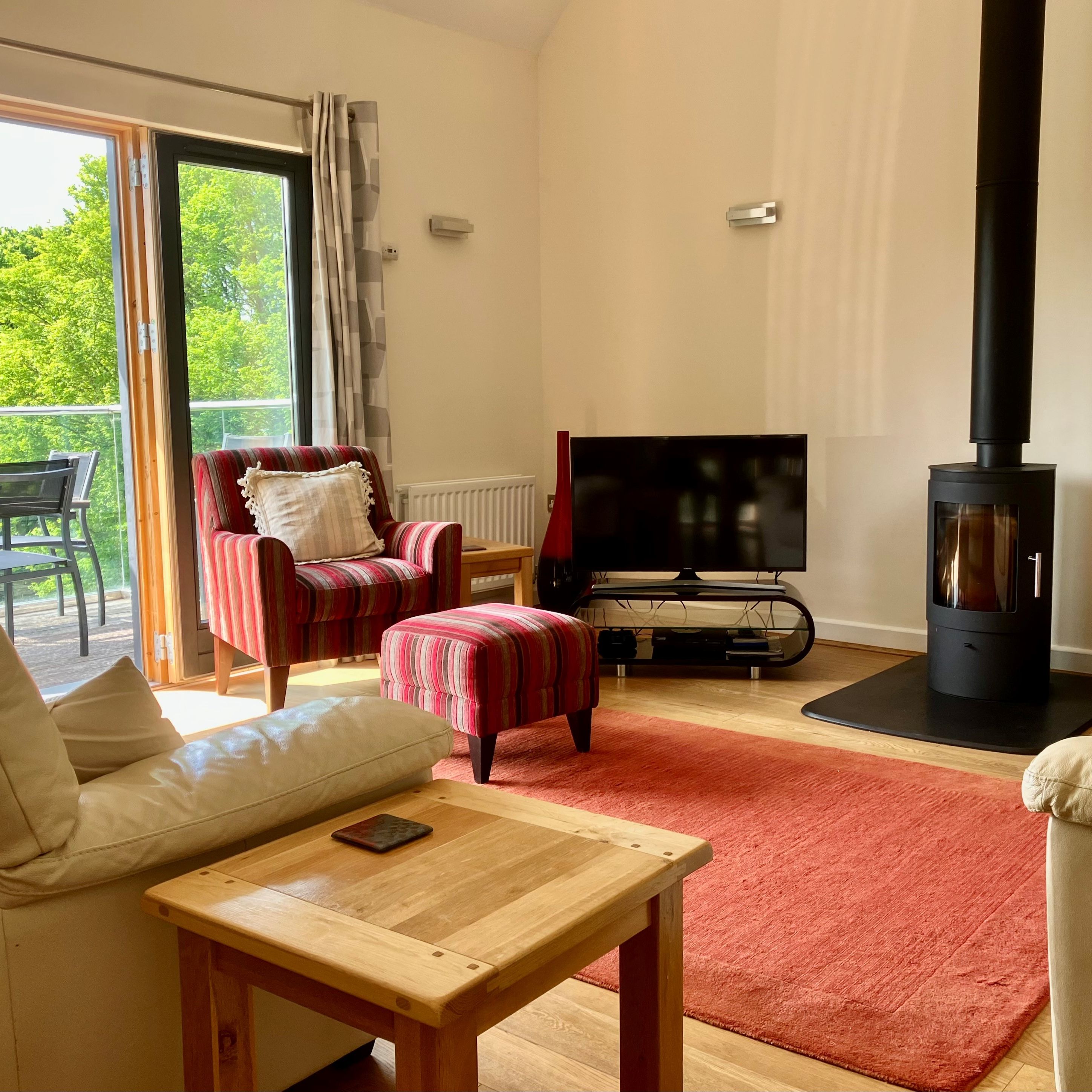 Cozy living room with a wood-burning stove, television, striped armchair, and large window opening to a balcony with outdoor seating.