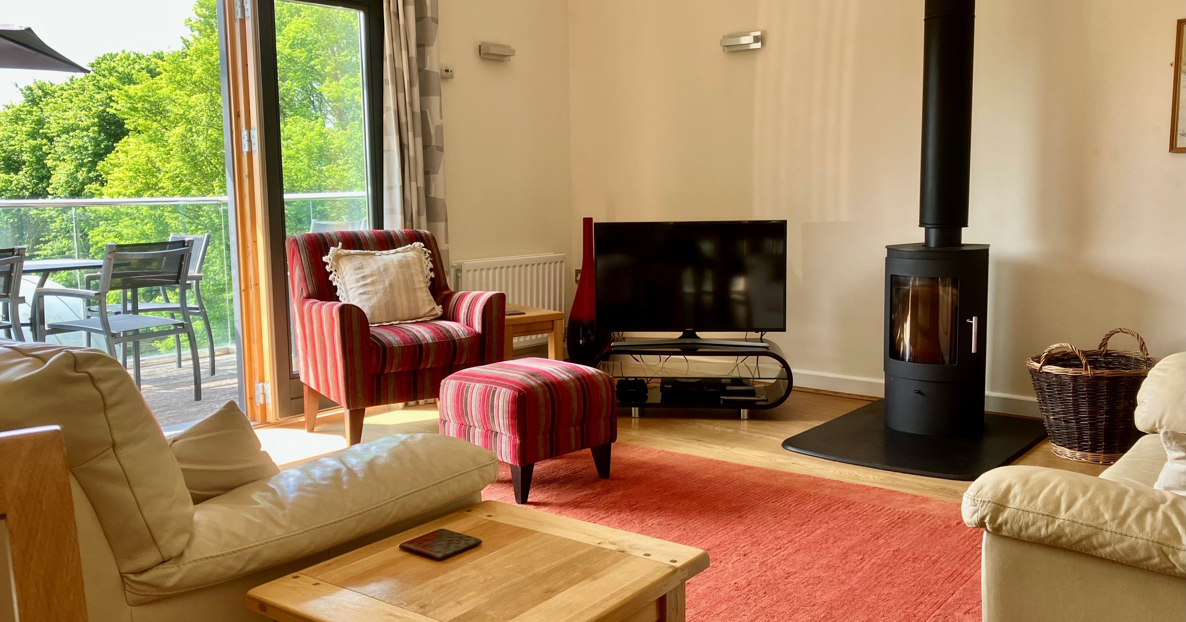 Cozy living room with a wood-burning stove, television, striped armchair, and large window opening to a balcony with outdoor seating.