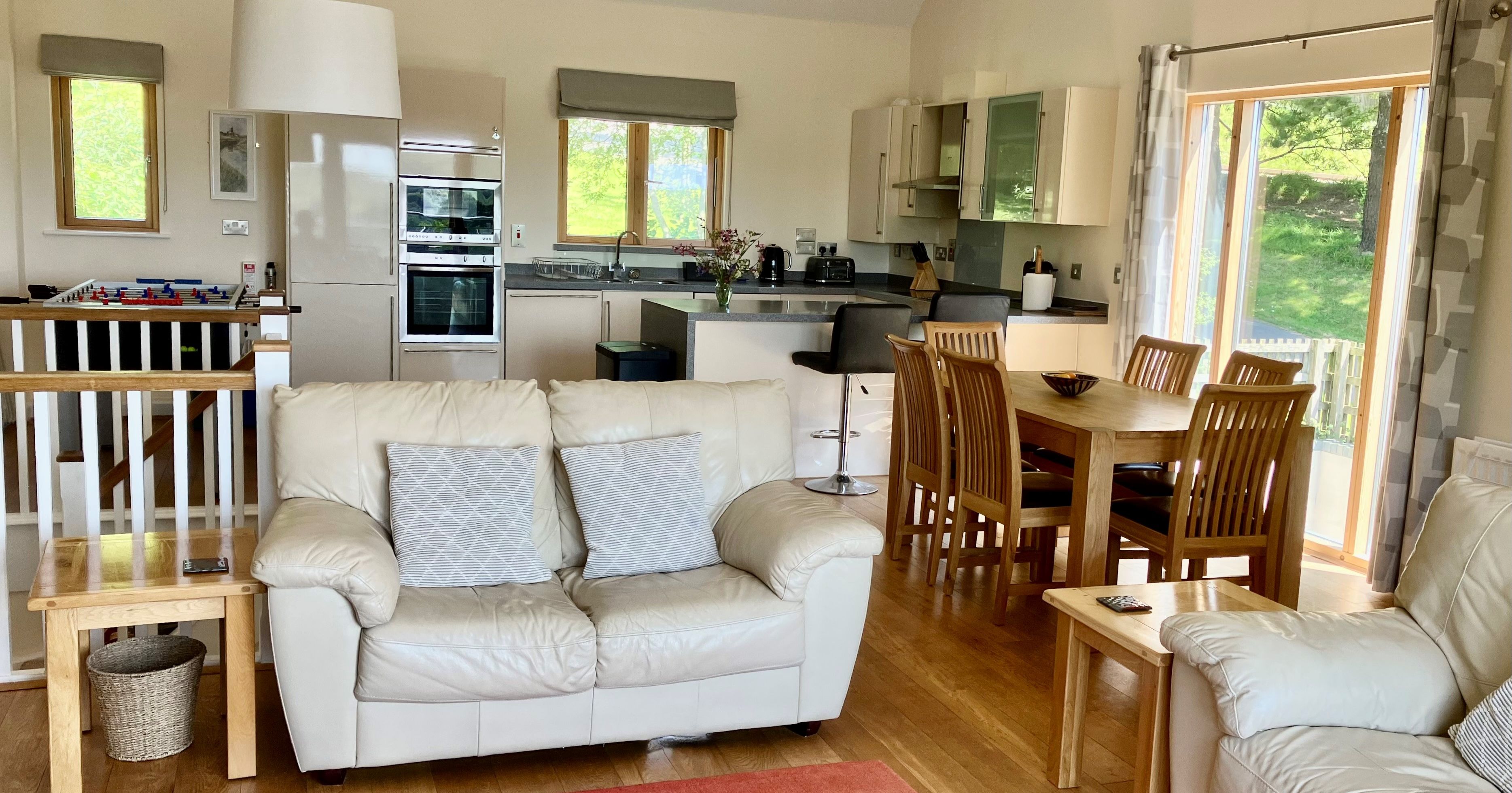 Modern open-plan living room and kitchen with white sofas, wooden furniture, and large windows.