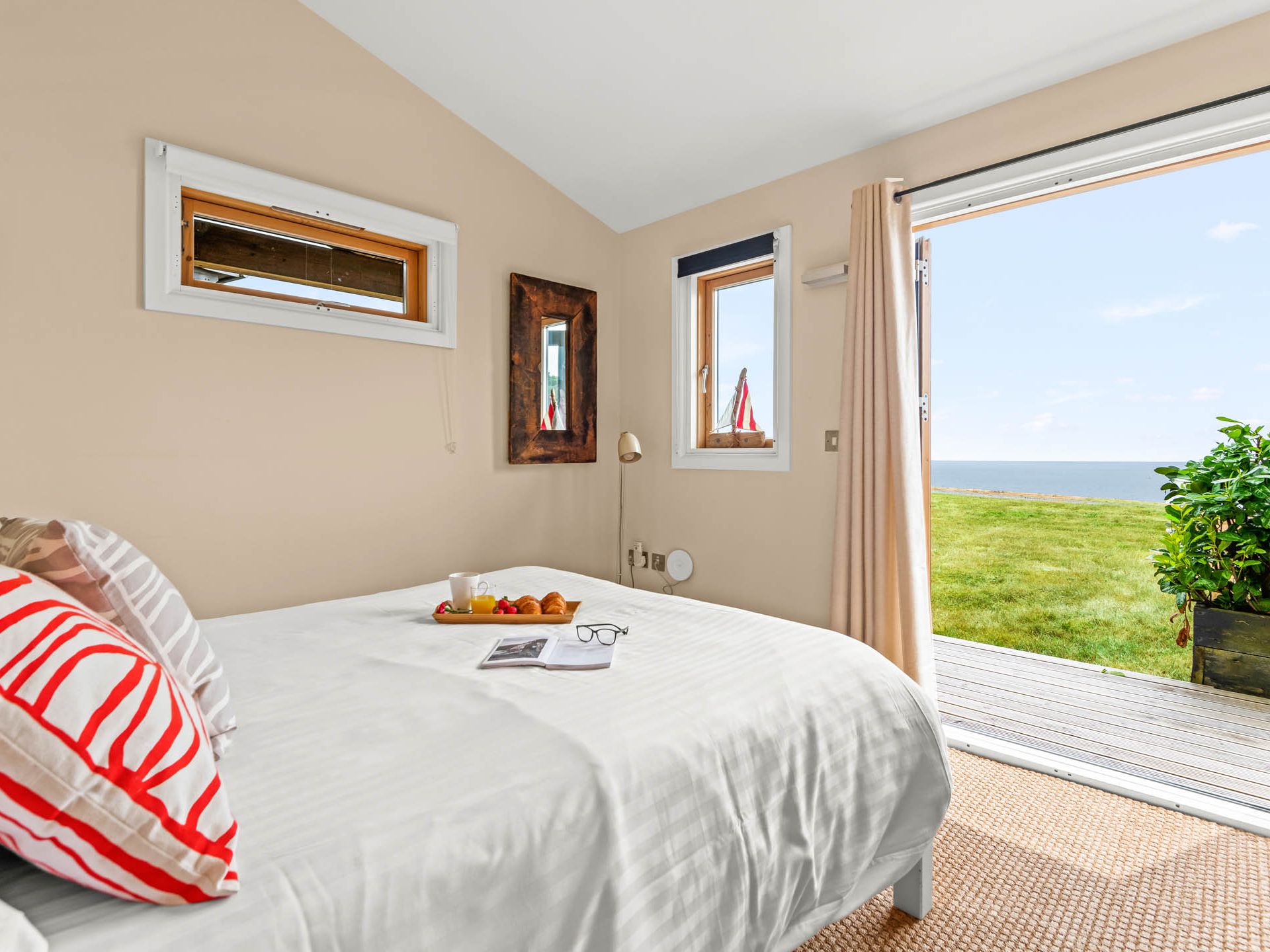 Bright bedroom with white bedding and red striped cushions, open door to a deck overlooking the sea