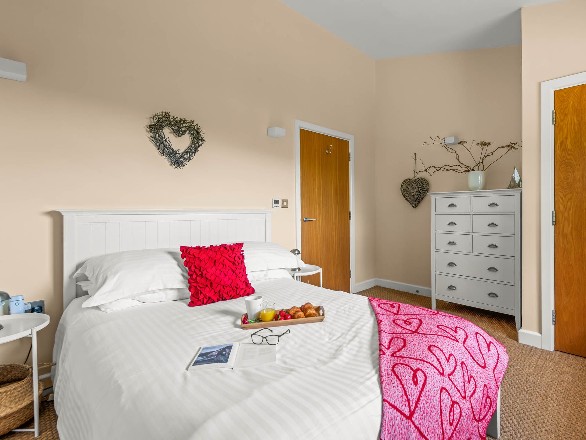 Bright bedroom with white bed, red cushion and pink throw, wooden doors and grey chest of drawers