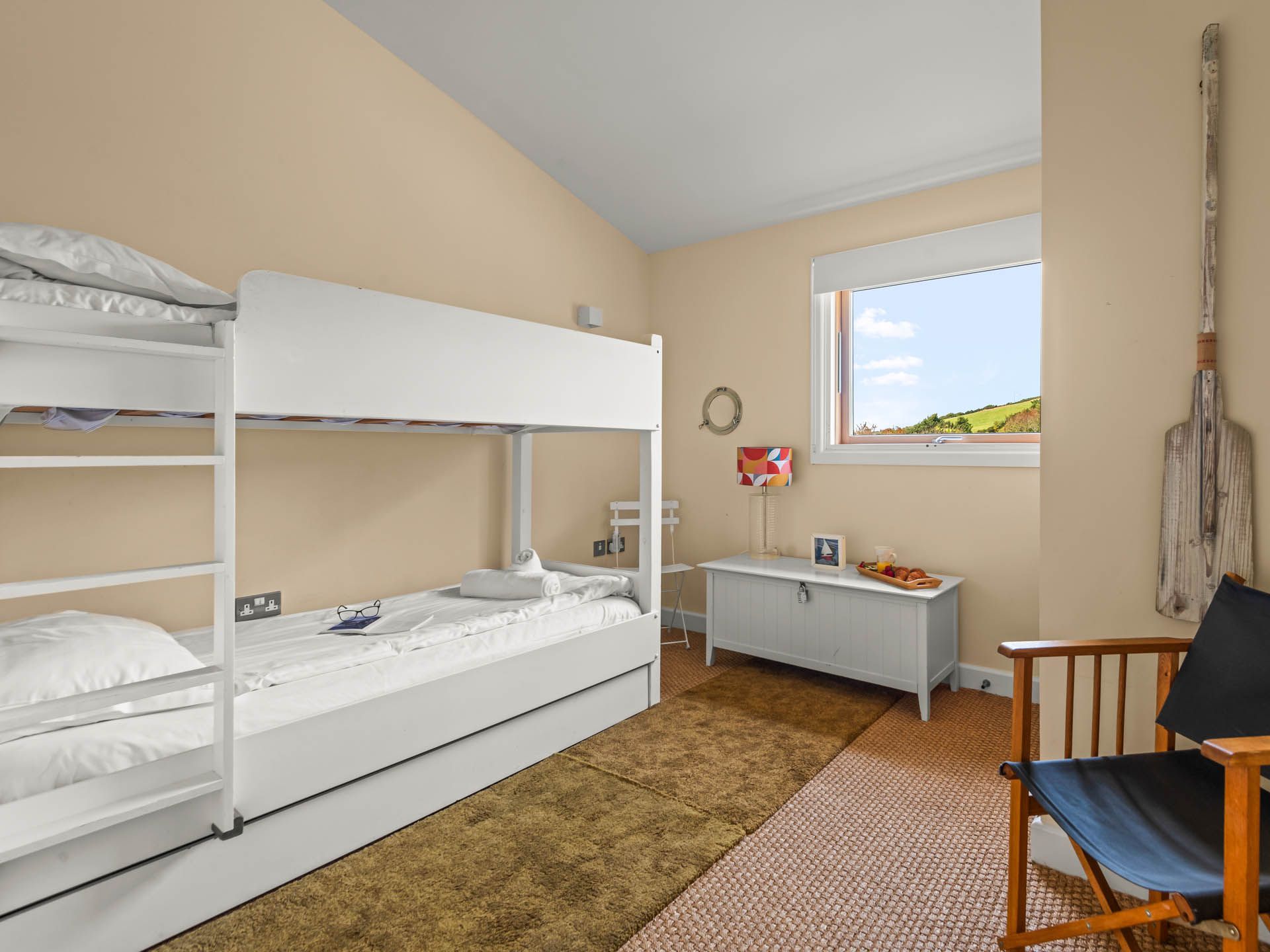 Bright bunk bedroom with white bunk beds, window view of green countryside, and wooden armchair