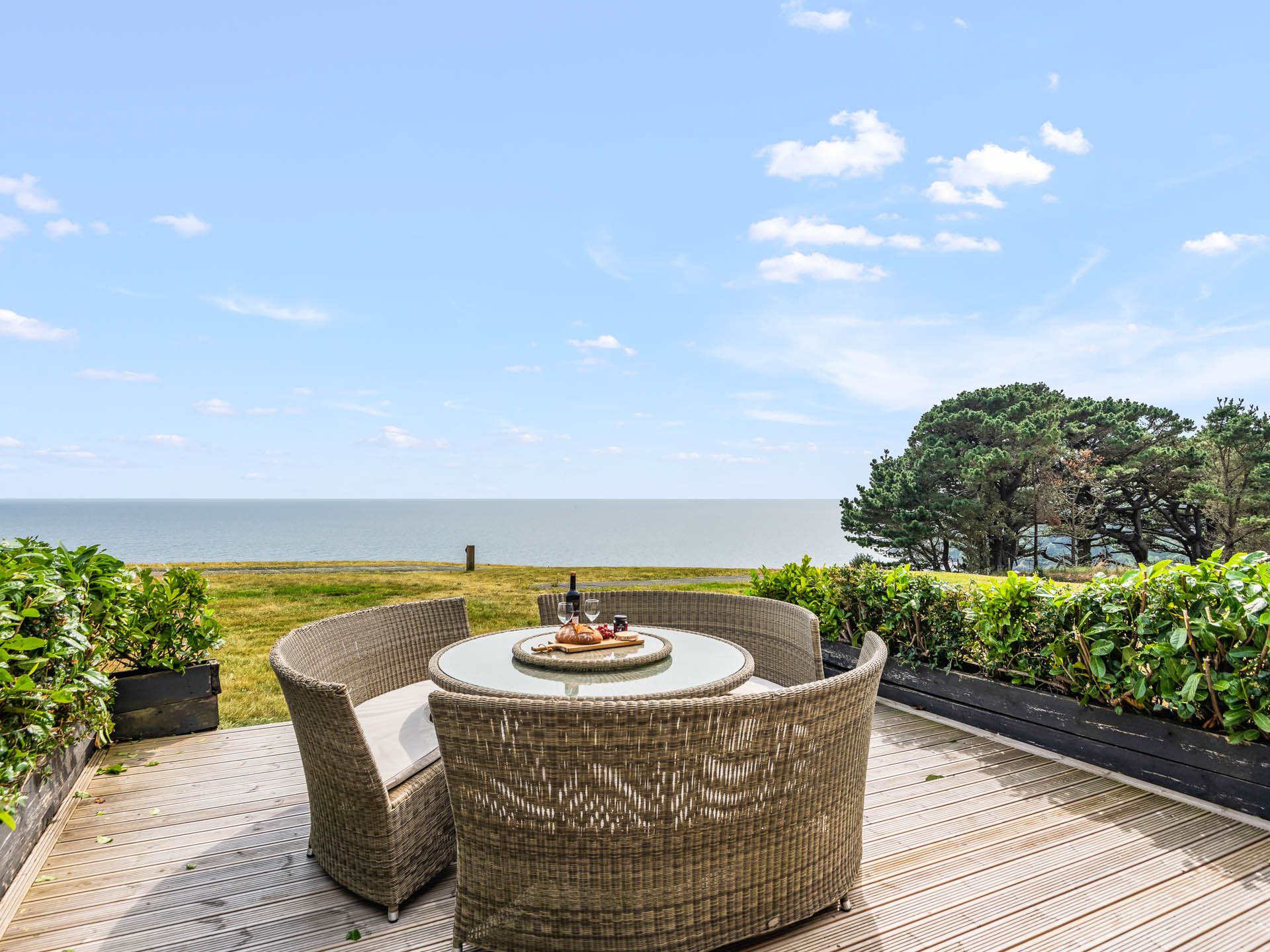 Outdoor wicker dining set on a wooden deck overlooking the sea under a blue sky