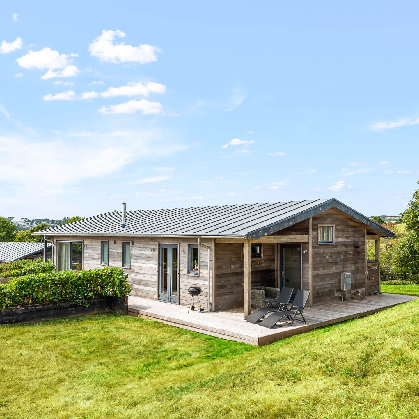 Modern wooden lodge with deck chairs on a raised terrace in a grassy hillside setting under a blue sky