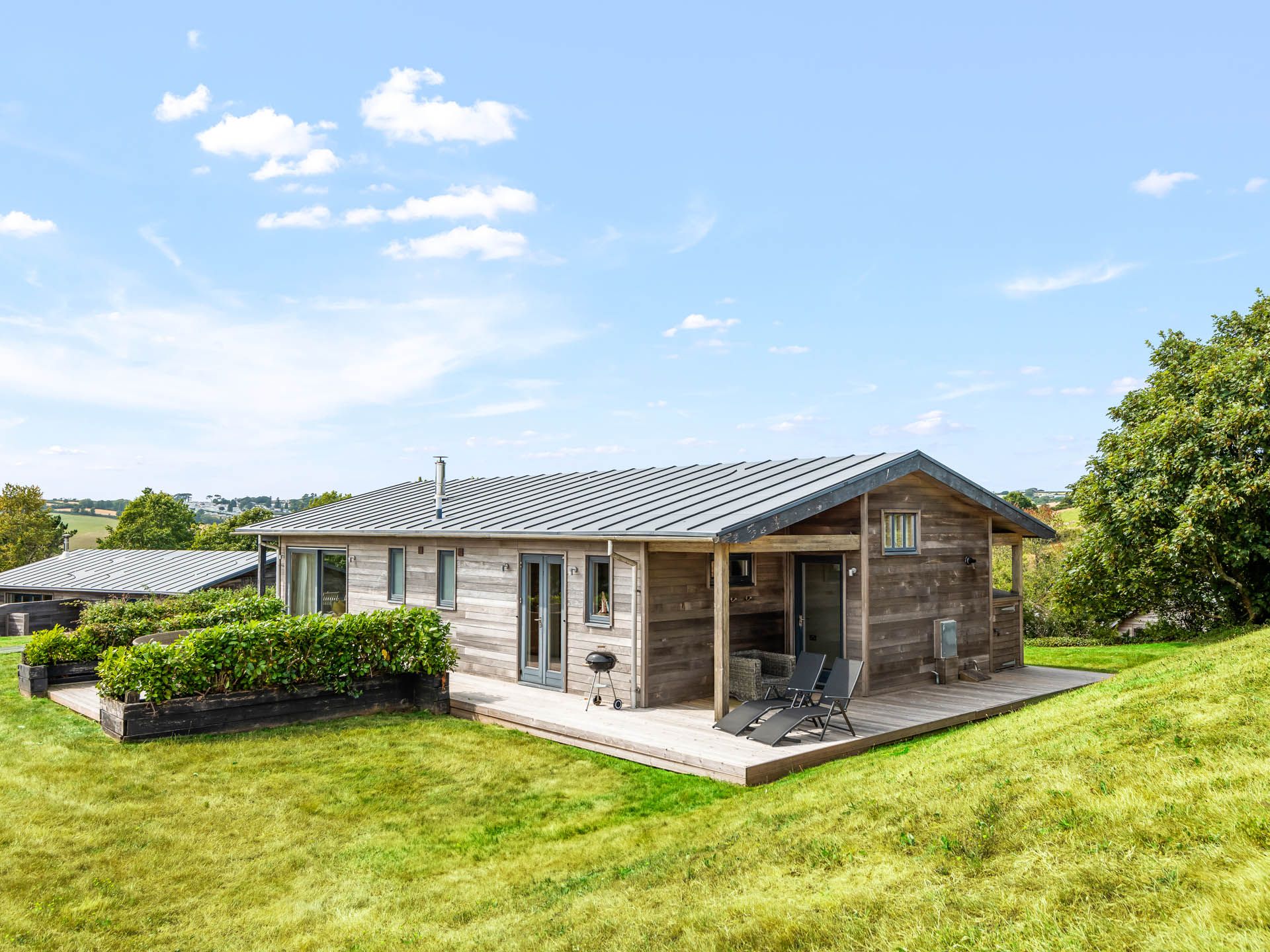Modern wooden lodge with deck chairs on a raised terrace in a grassy hillside setting under a blue sky