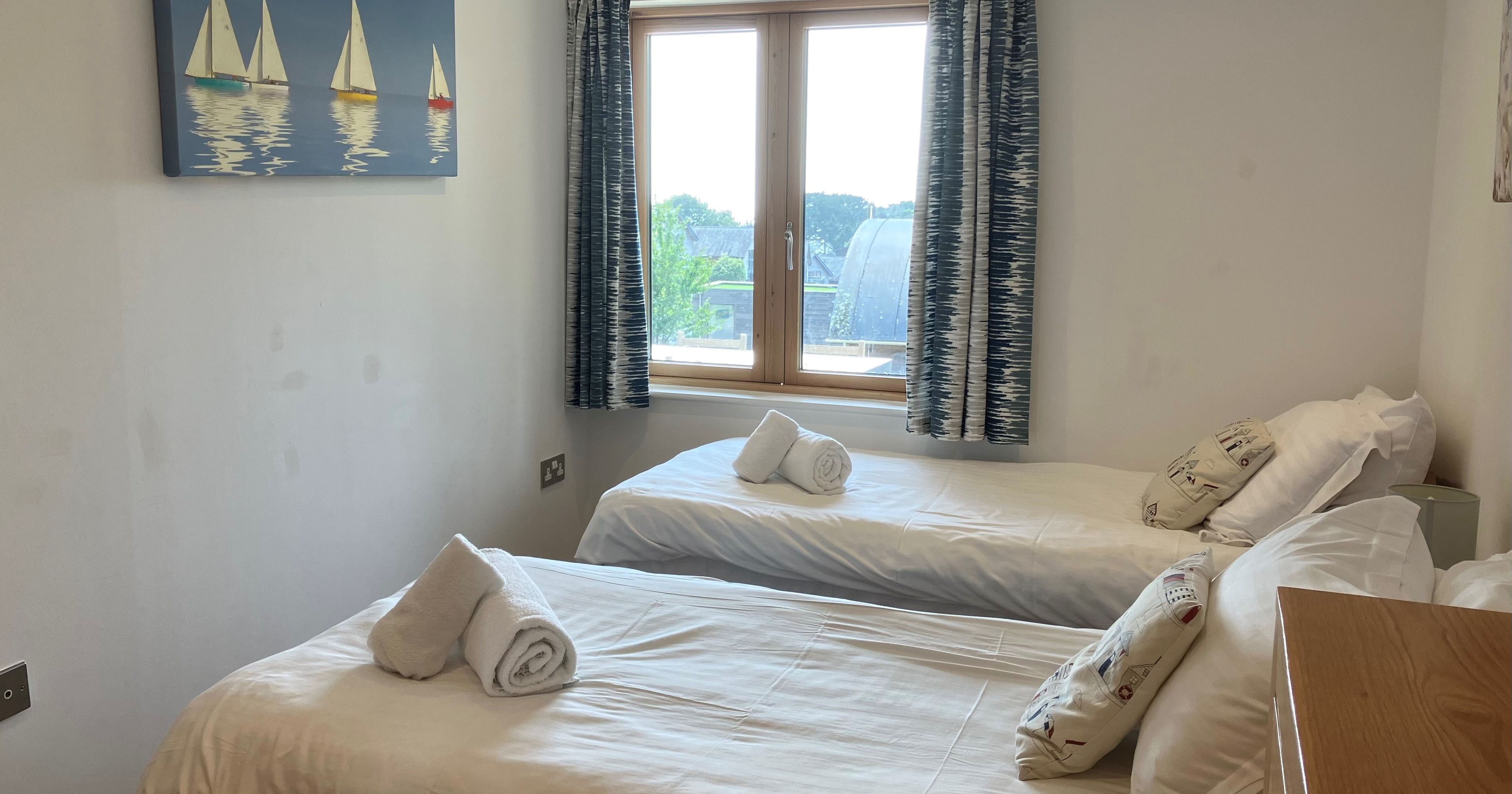 A small bedroom with two single beds, each topped with rolled white towels, and a window with patterned curtains.