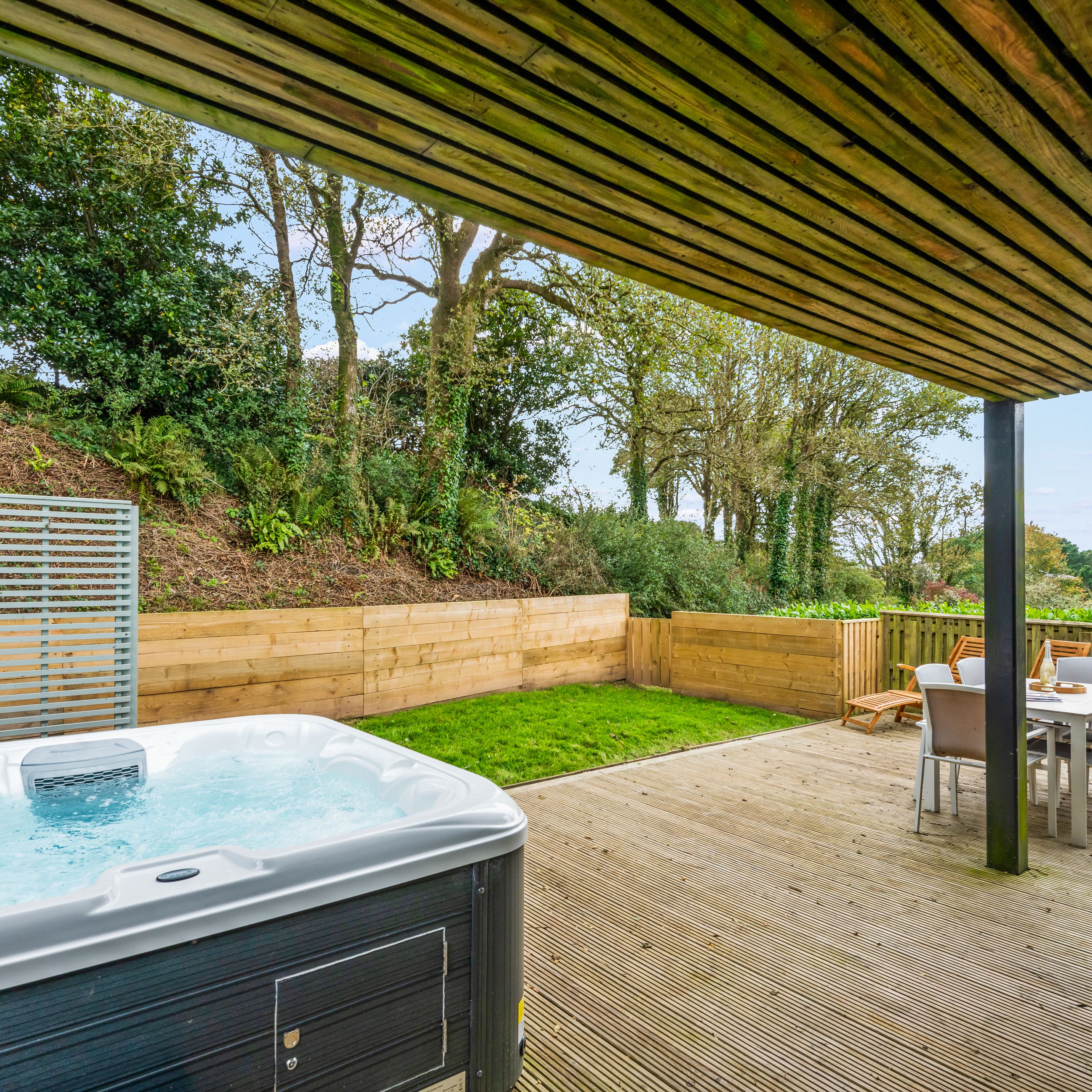 Covered wooden patio with a hot tub, outdoor dining table and chairs, and a view of a landscaped garden with trees and a wooden fence.