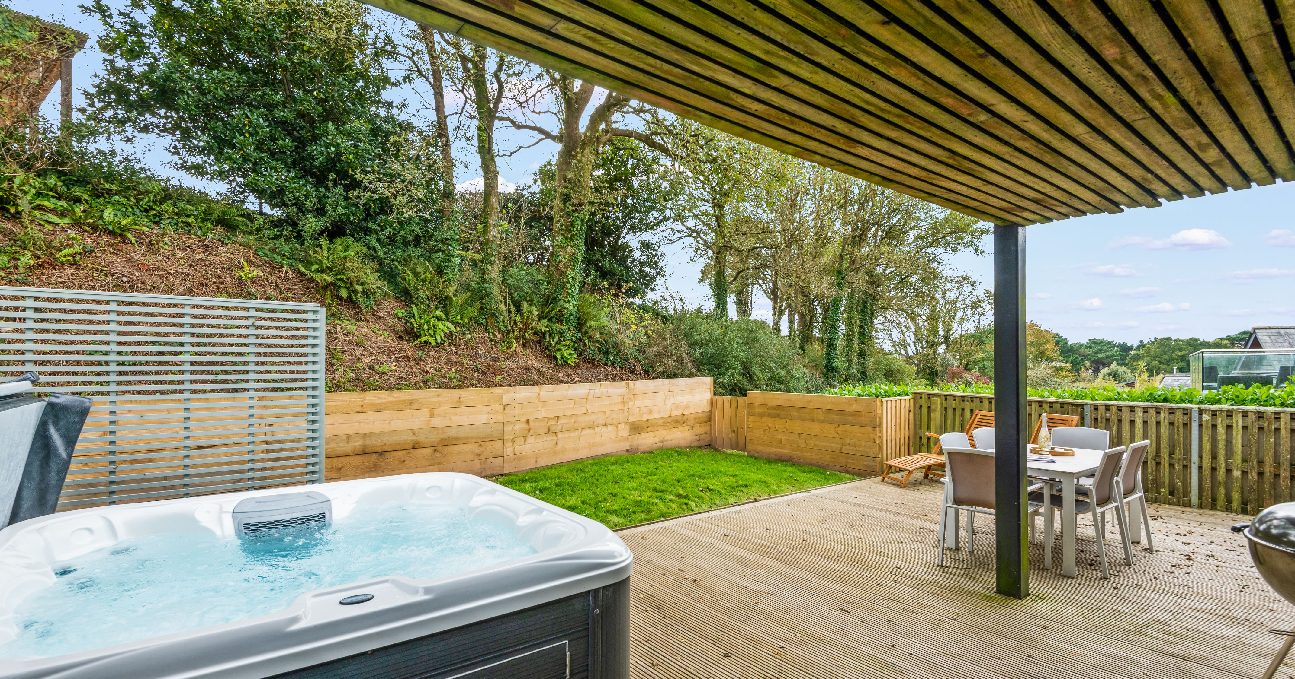 Covered wooden patio with a hot tub, outdoor dining table and chairs, and a view of a landscaped garden with trees and a wooden fence.