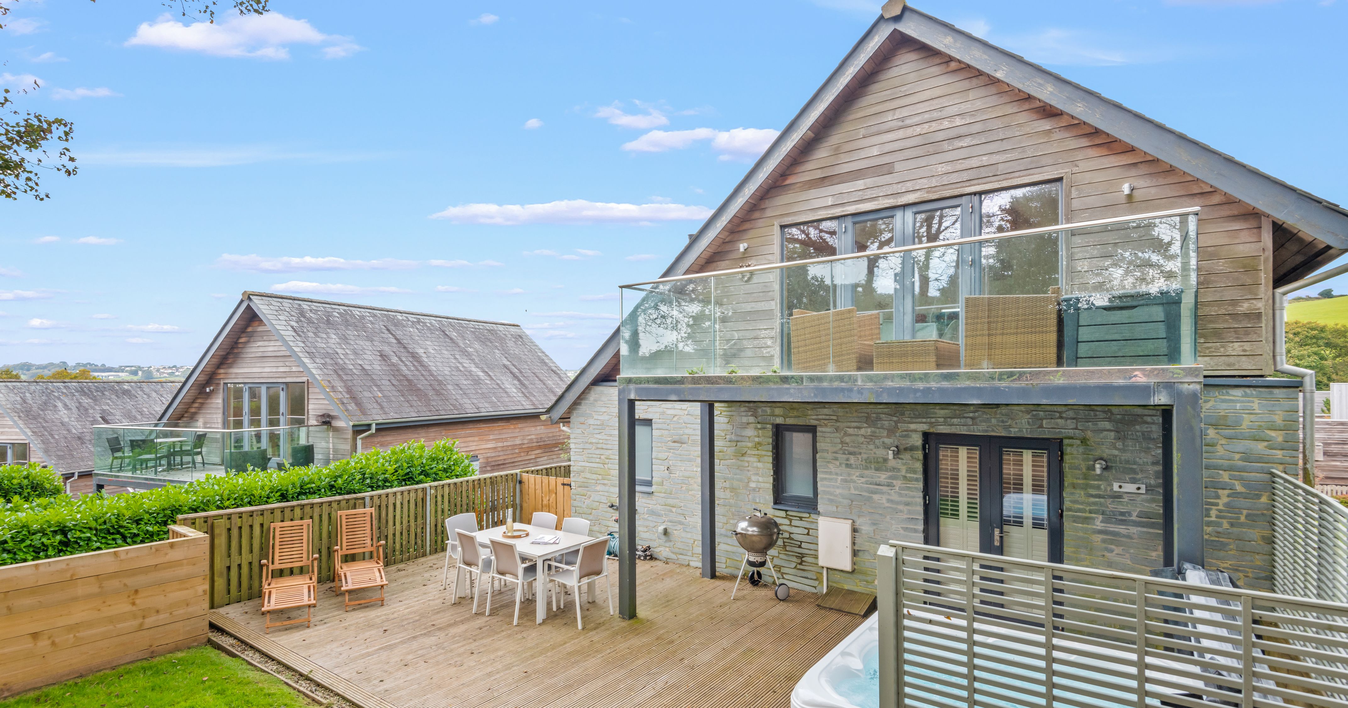 Modern two-story house with a spacious deck, outdoor dining area, lounge chairs, hot tub, and glass balcony.