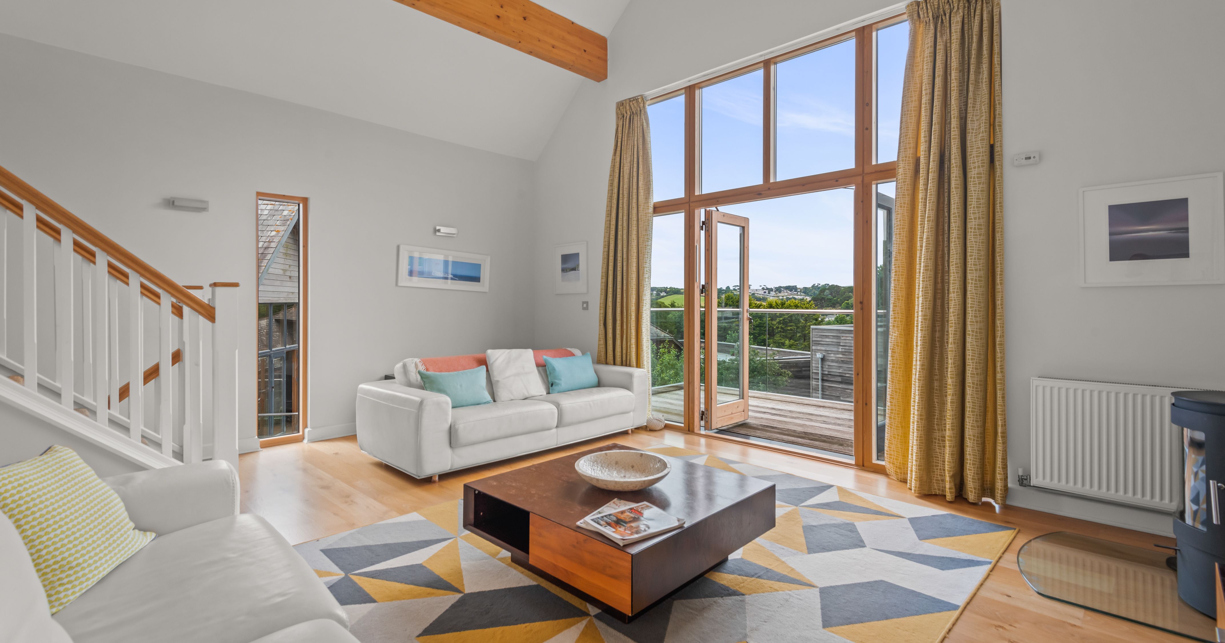 Modern living room with large windows, white sofas, geometric rug, and view of outdoor deck.