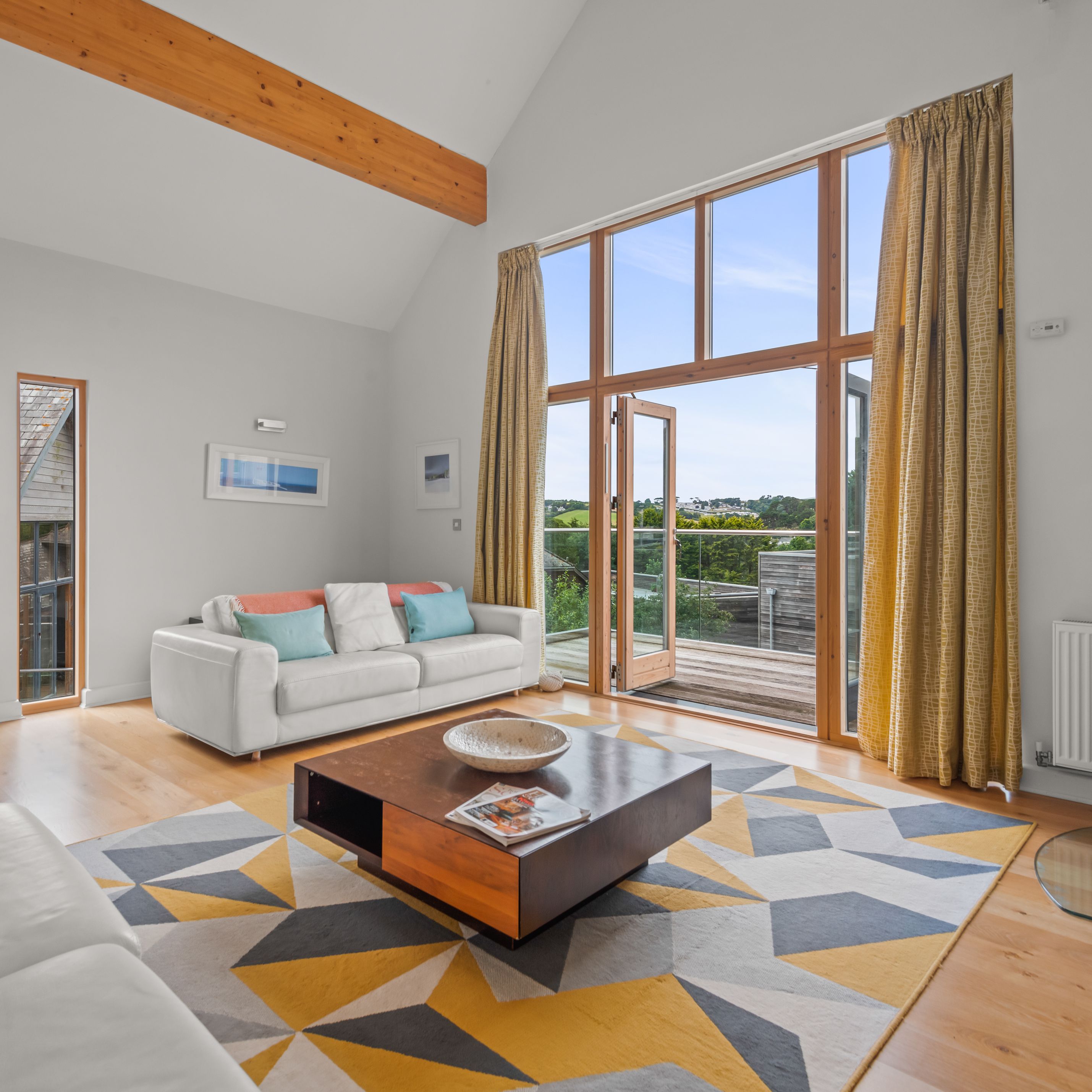 Modern living room with large windows, white sofas, geometric rug, and view of outdoor deck.