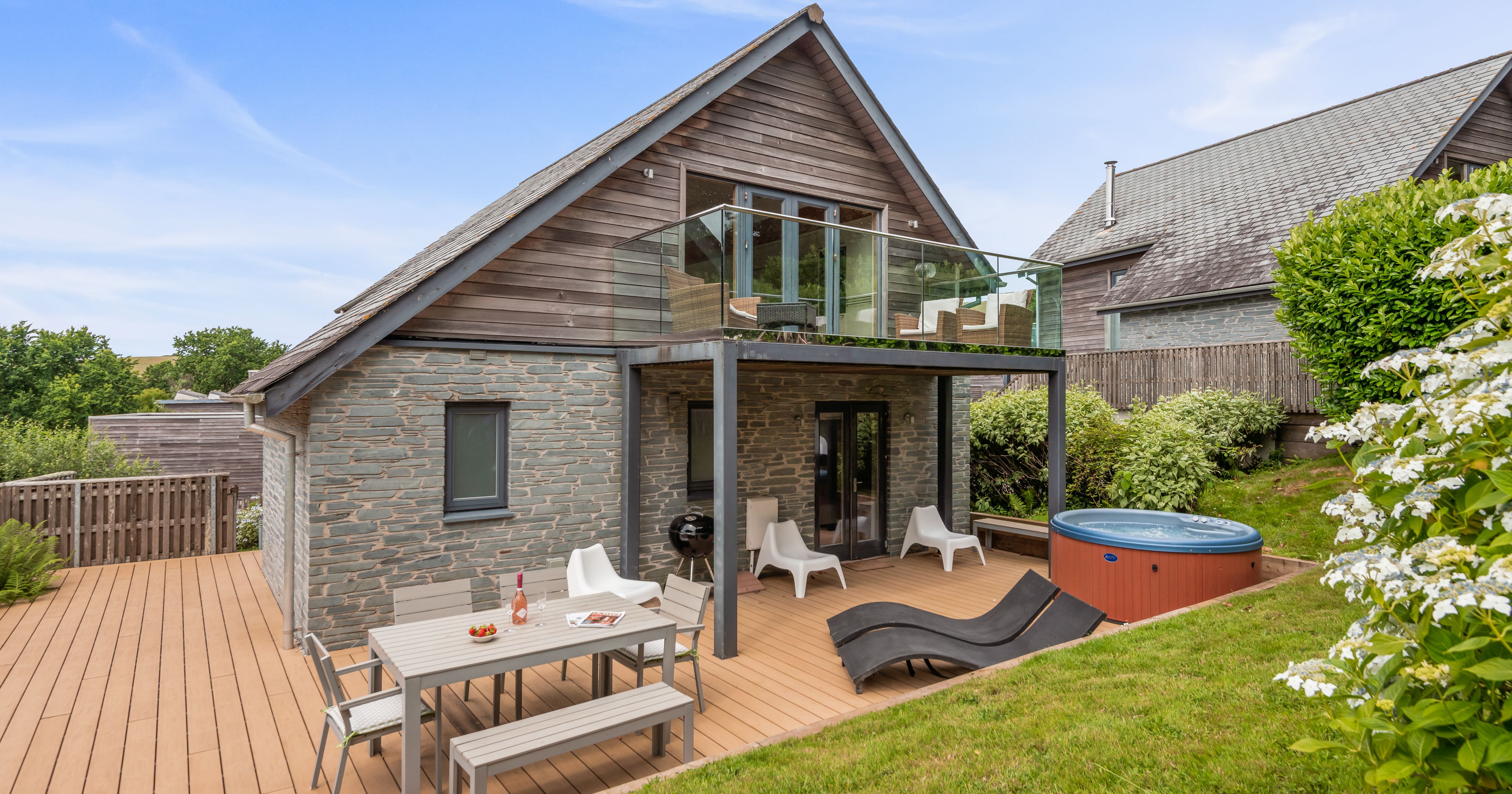 Modern house with stone and wood exterior, upper balcony with glass railing, patio furniture, hot tub, and landscaped garden