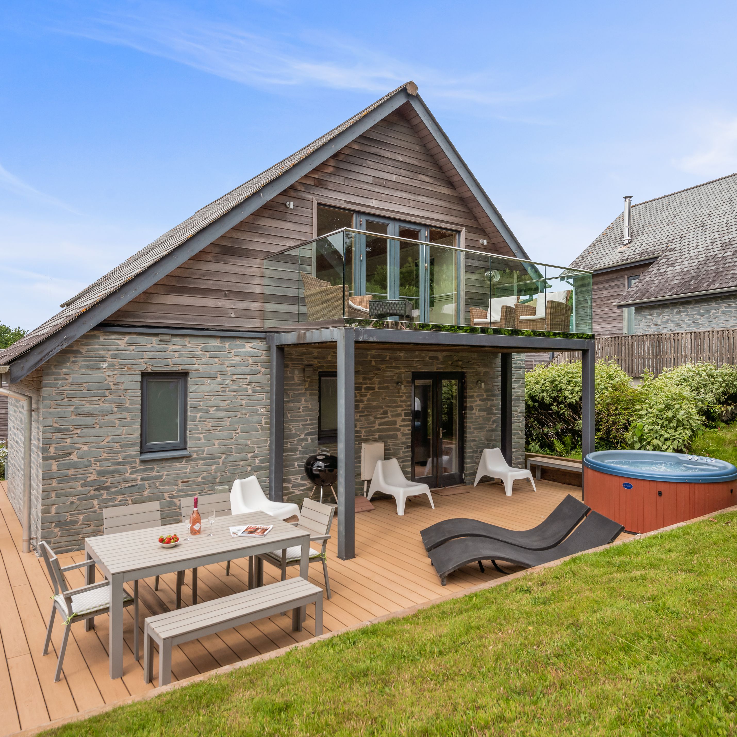 Modern house with stone and wood exterior, upper balcony with glass railing, patio furniture, hot tub, and landscaped garden