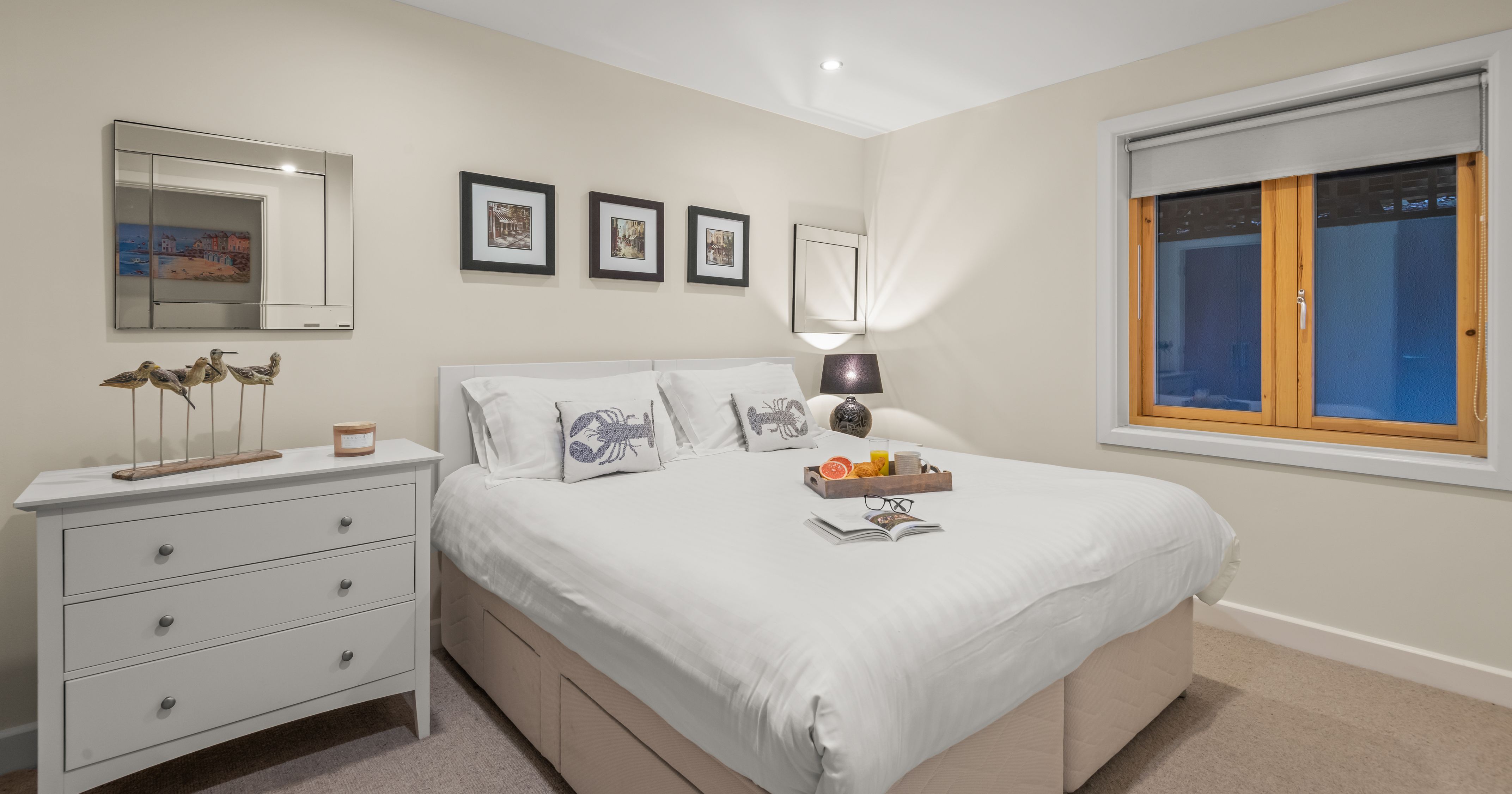 Modern cozy bedroom with white bedding, artwork above the bed, and a window with wooden trim.
