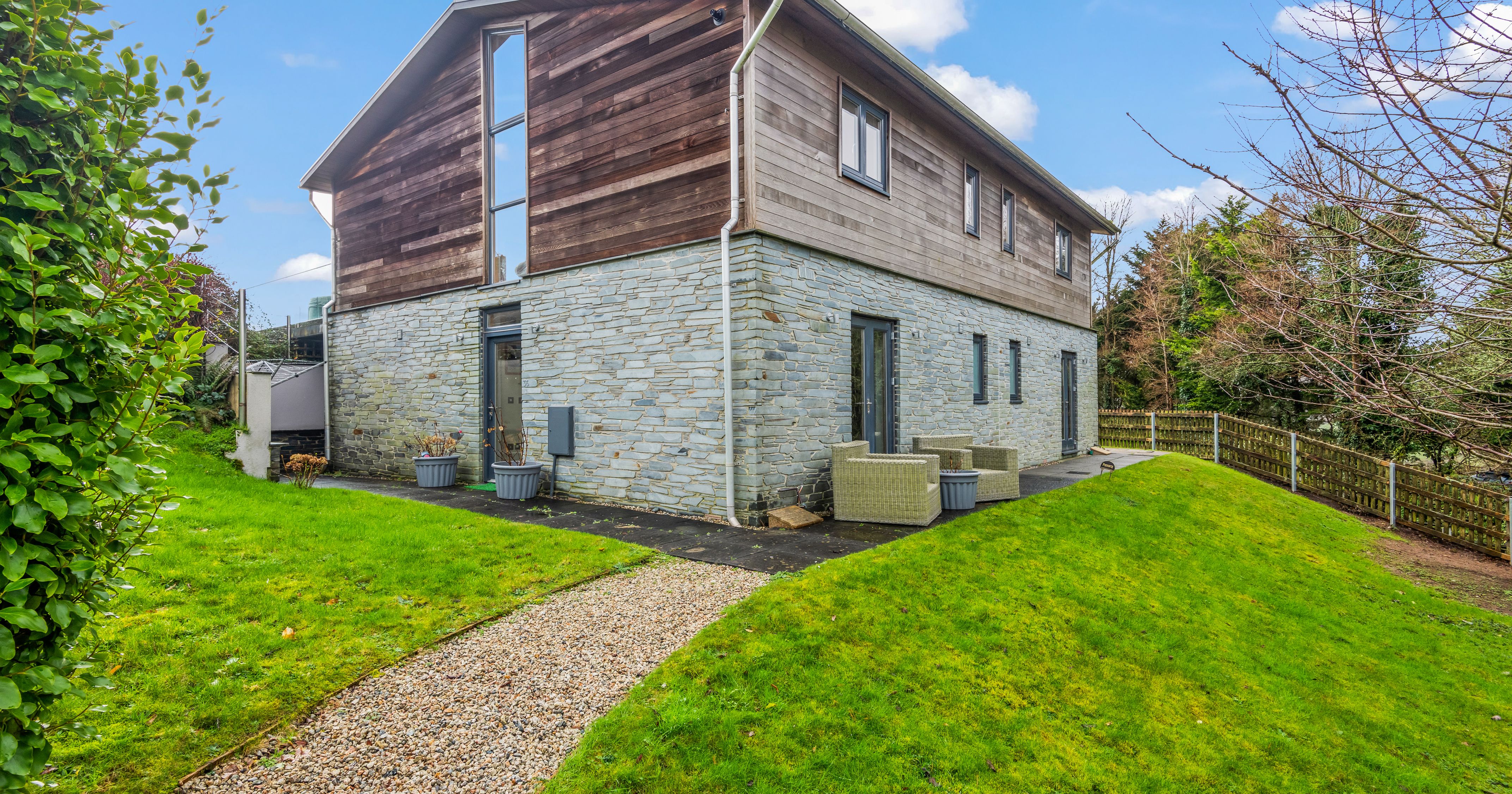 Modern two-story house with stone and wood exterior, surrounded by green lawn and outdoor patio furniture.