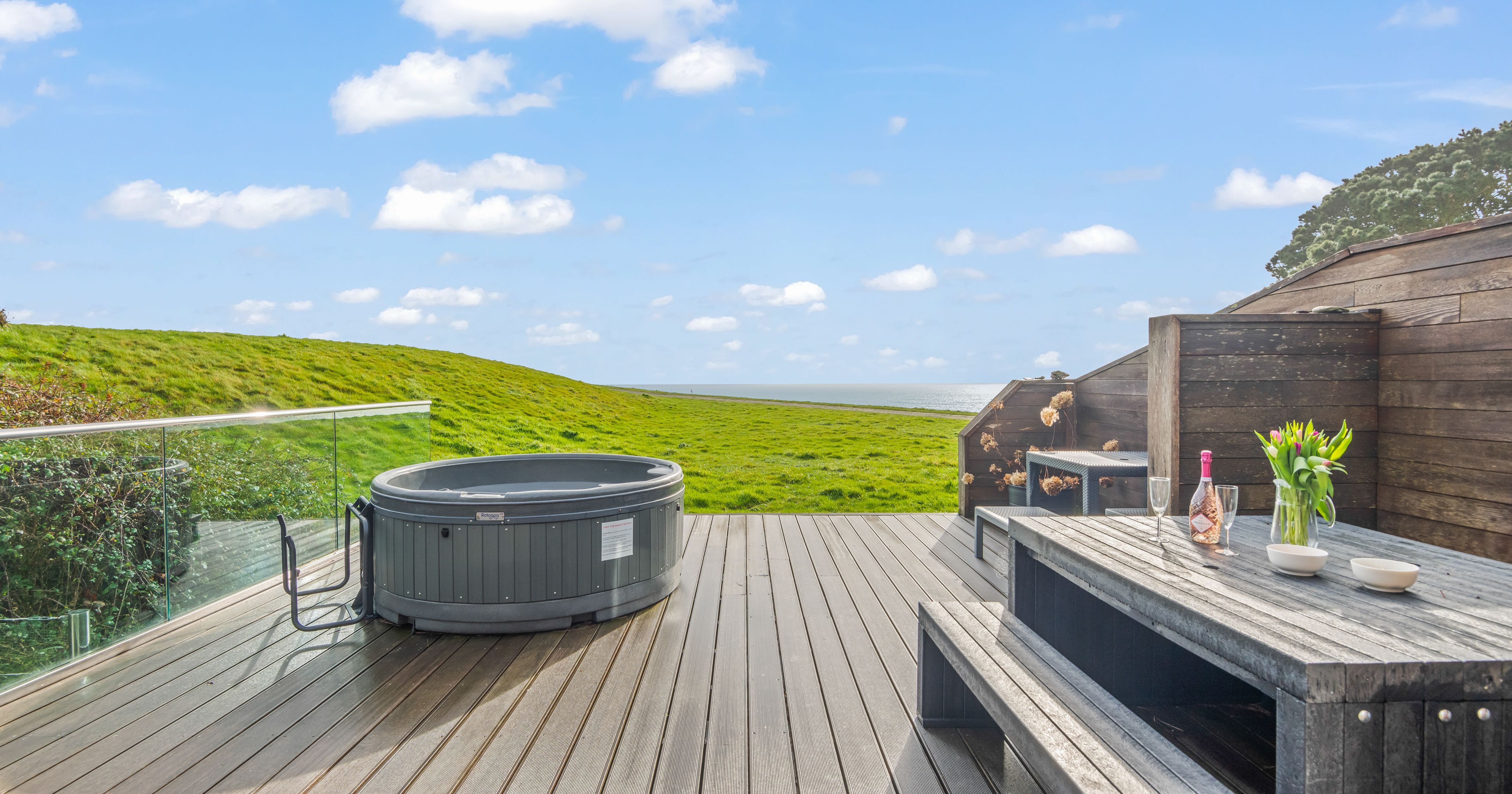 Modern outdoor deck with hot tub, glass railing, and picnic table overlooking grassy hills.
