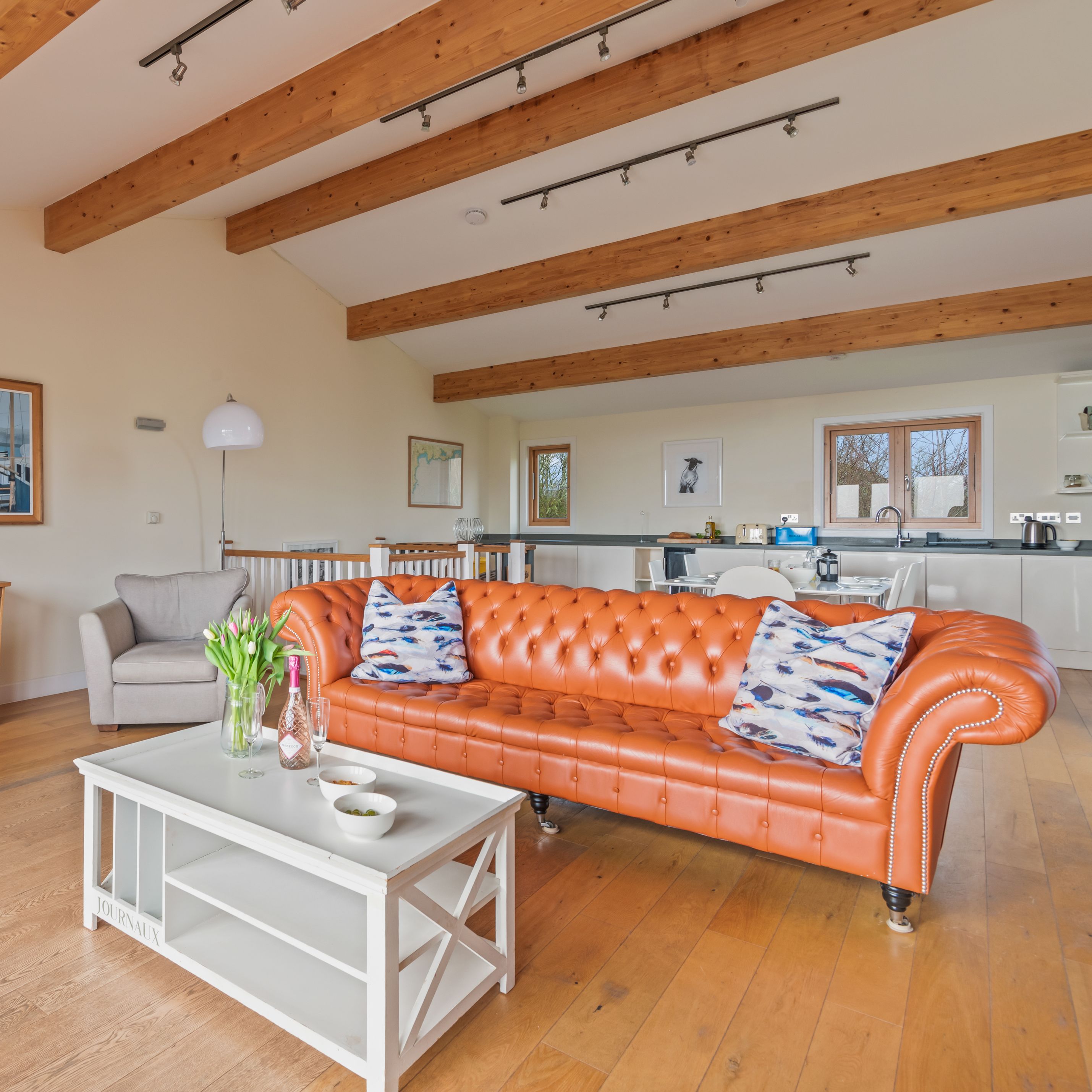 Modern open-plan living room with orange leather sofa, wooden beams, and kitchen area in background