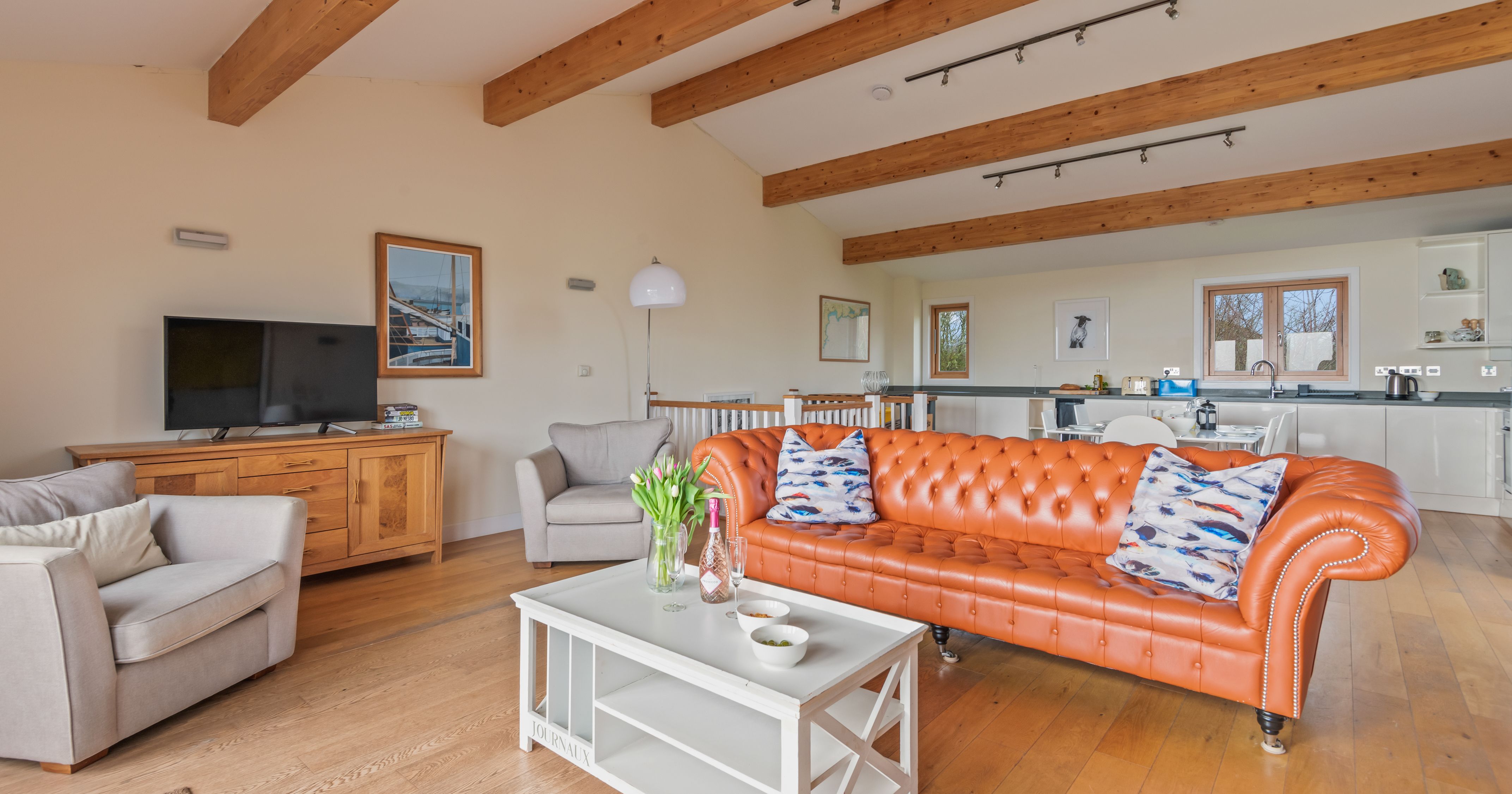 Modern open-plan living room with orange leather sofa, wooden beams, and kitchen area in background