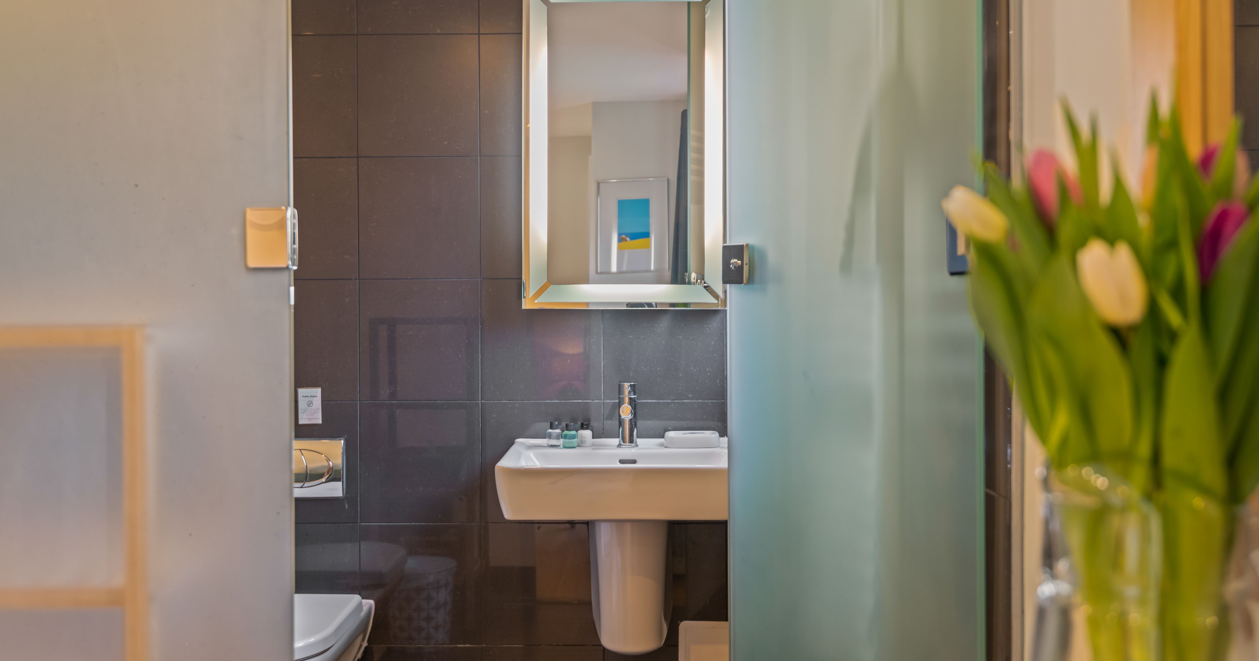 Modern bathroom with a white sink, large mirror, dark tiled wall, frosted glass door, a toilet partially visible, and a vase with colorful tulips in the foreground.