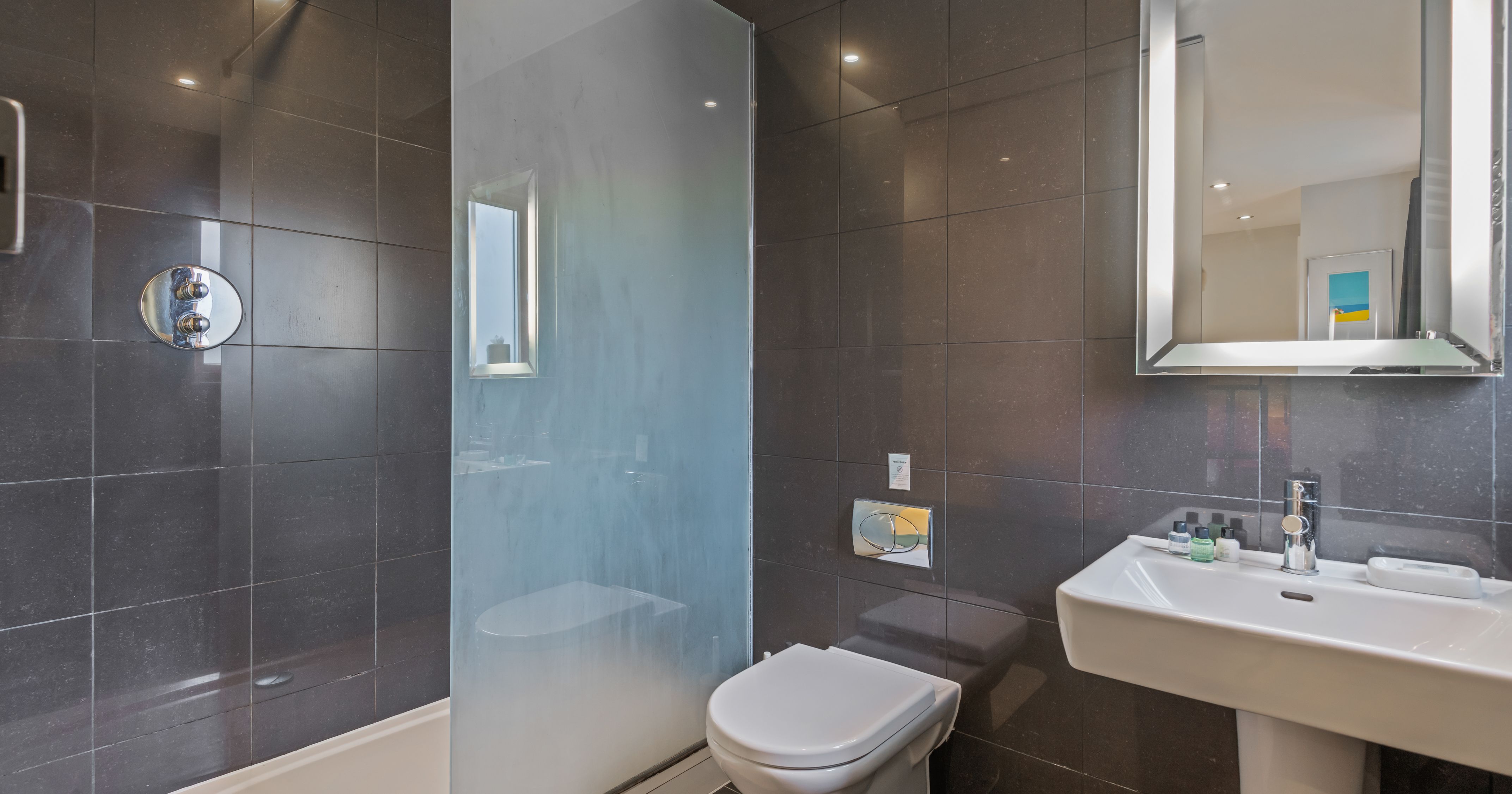 Modern bathroom with dark tiled walls, a walk-in shower with frosted glass partition, wall-mounted toilet, and a white sink with a mirror above.