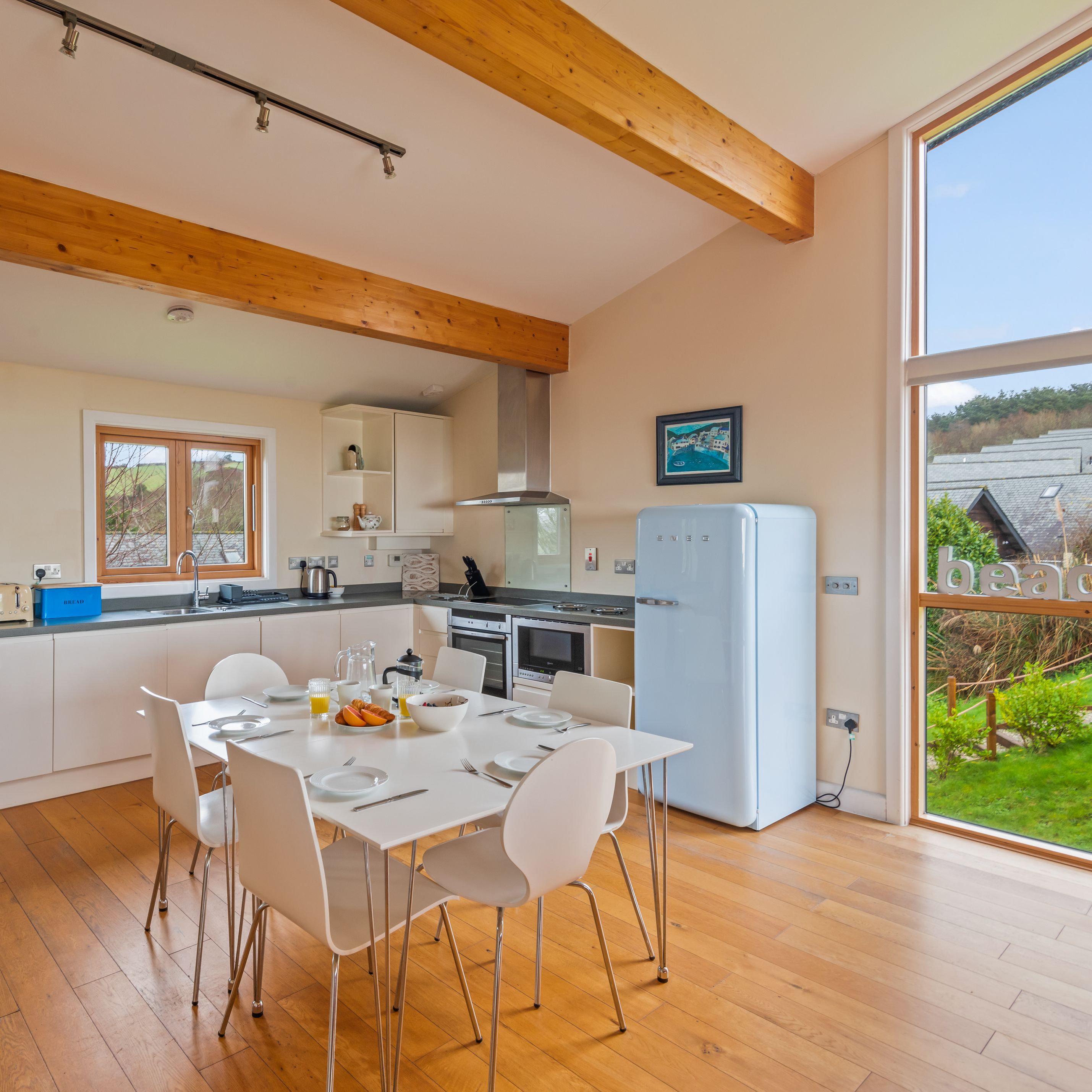 Modern kitchen with white cabinets, dining table, wooden floors, and large window overlooking a garden