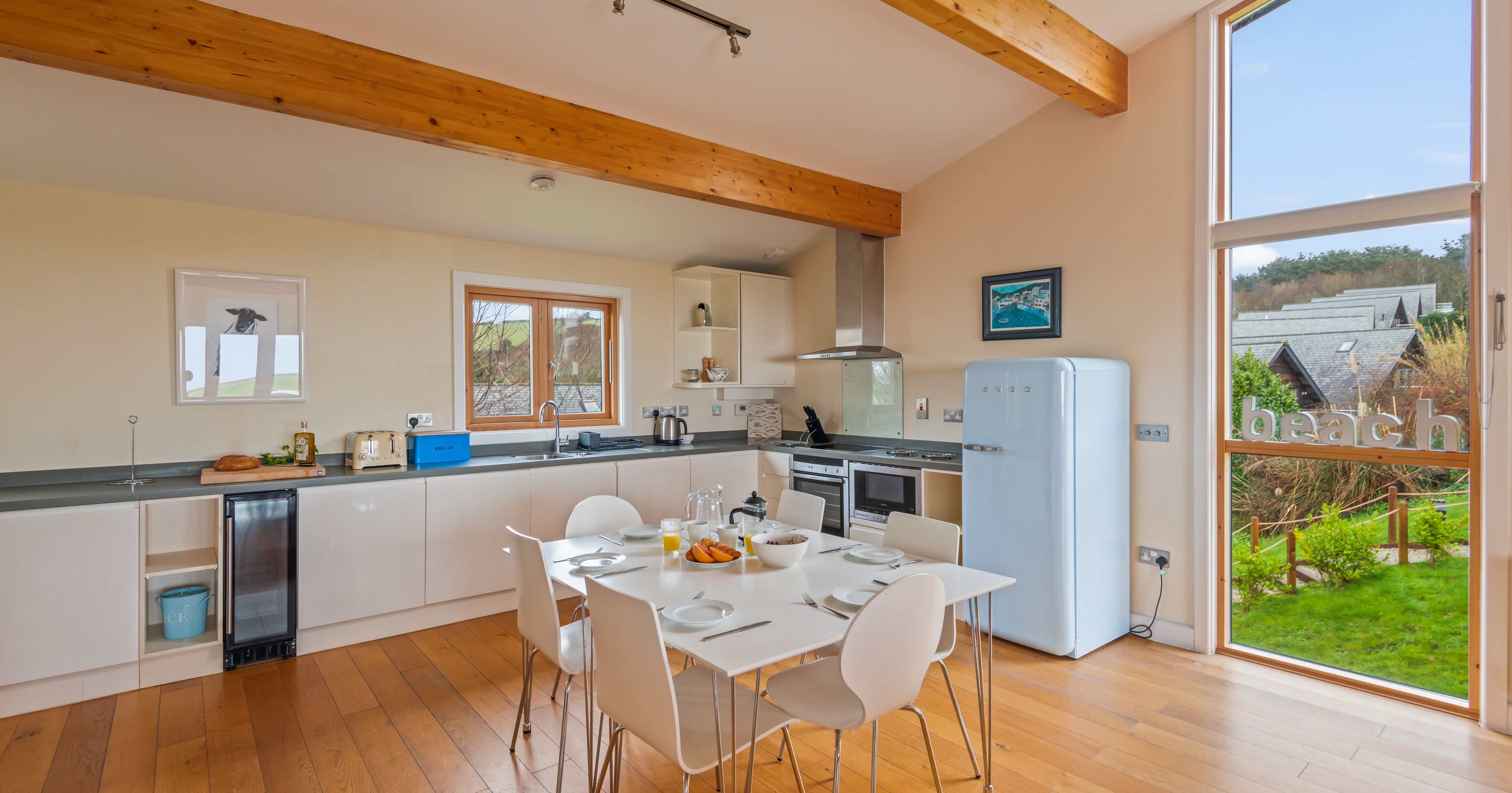 Modern kitchen with white cabinets, dining table, wooden floors, and large window overlooking a garden