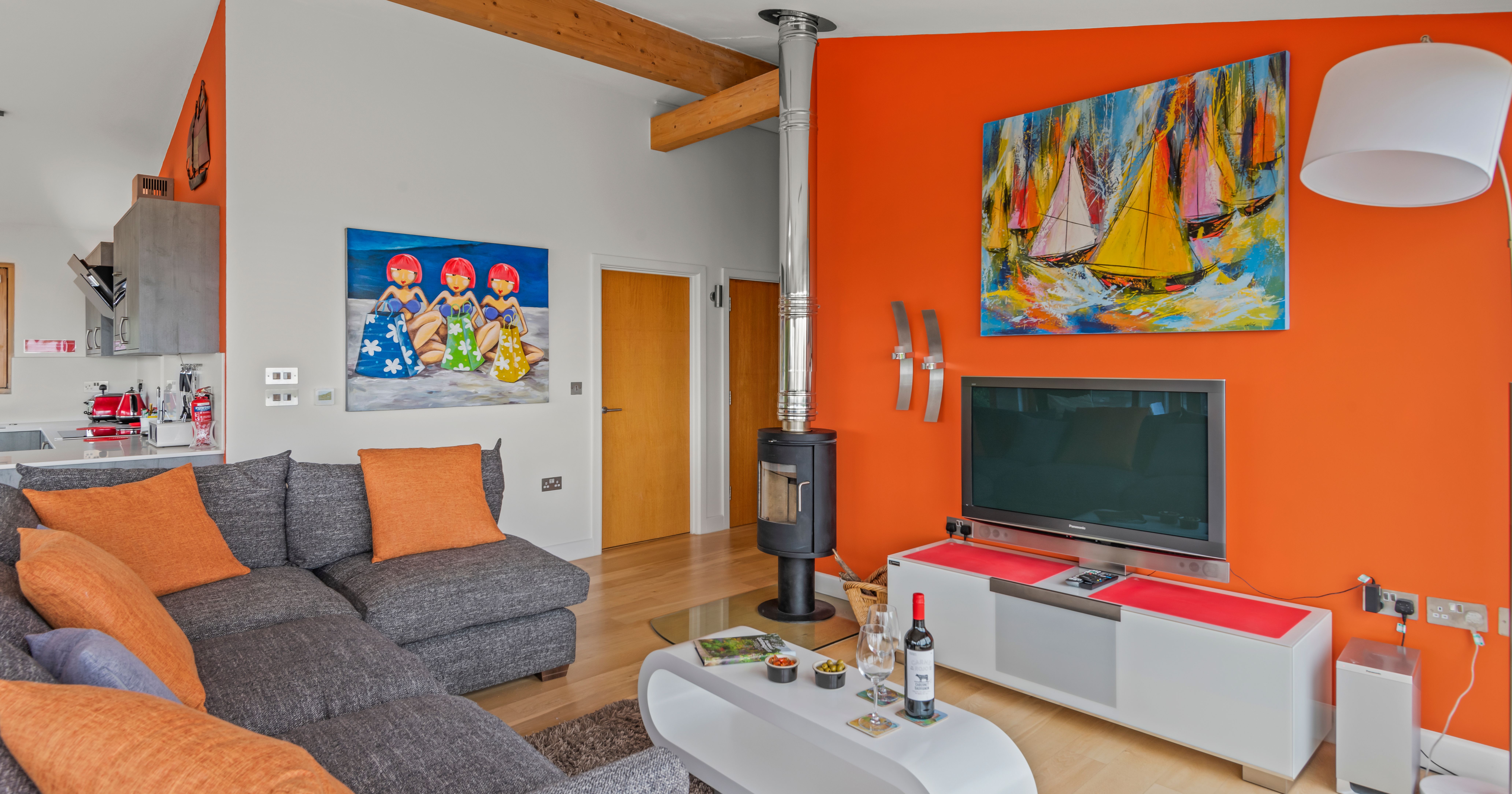 Modern living room with orange accent wall, grey sectional sofa, colorful paintings, and a TV.