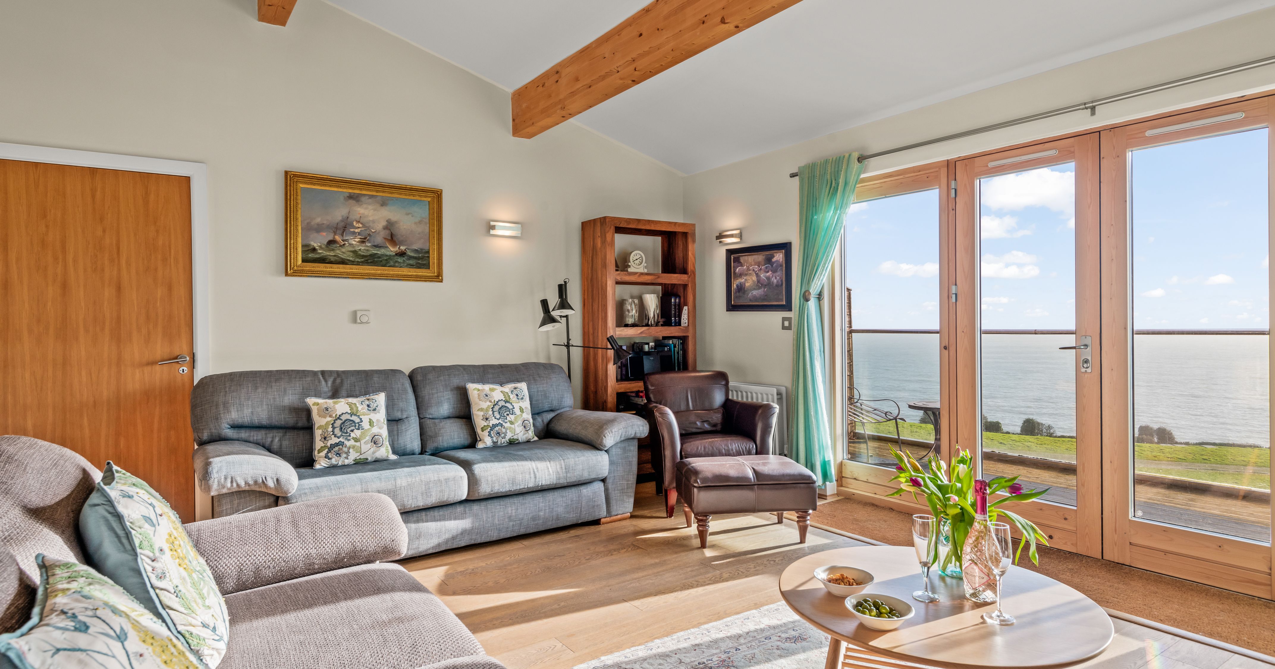 Cozy living room with large windows overlooking the sea, featuring comfortable sofas, a wooden coffee table, and decorative cushions.