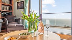 A cozy living room with a leather armchair, a round table set with a bottle of rose prosecco, two glasses, bowls of snacks, a vase of tulips, and a scenic ocean view through glass doors.