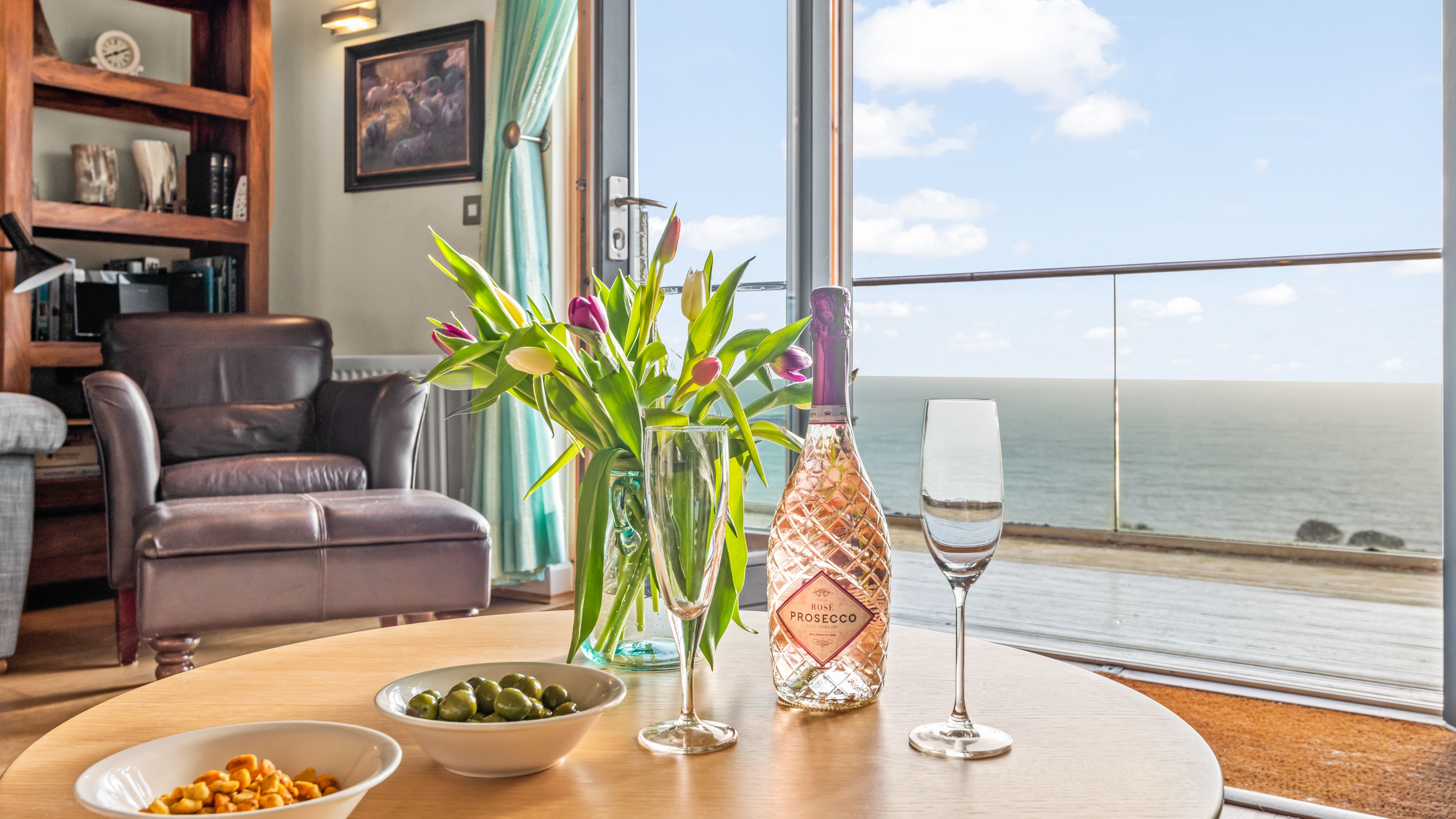 A cozy living room with a leather armchair, a round table set with a bottle of rose prosecco, two glasses, bowls of snacks, a vase of tulips, and a scenic ocean view through glass doors.