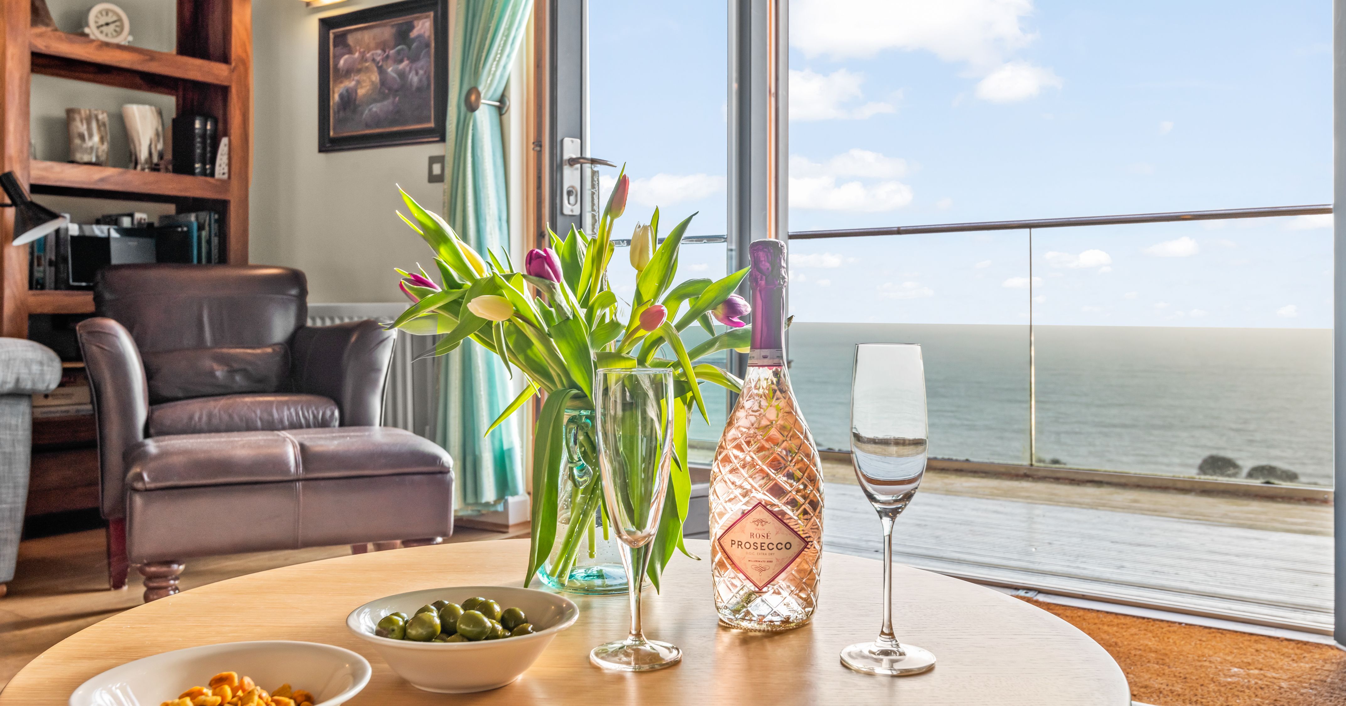 A cozy living room with a leather armchair, a round table set with a bottle of rose prosecco, two glasses, bowls of snacks, a vase of tulips, and a scenic ocean view through glass doors.