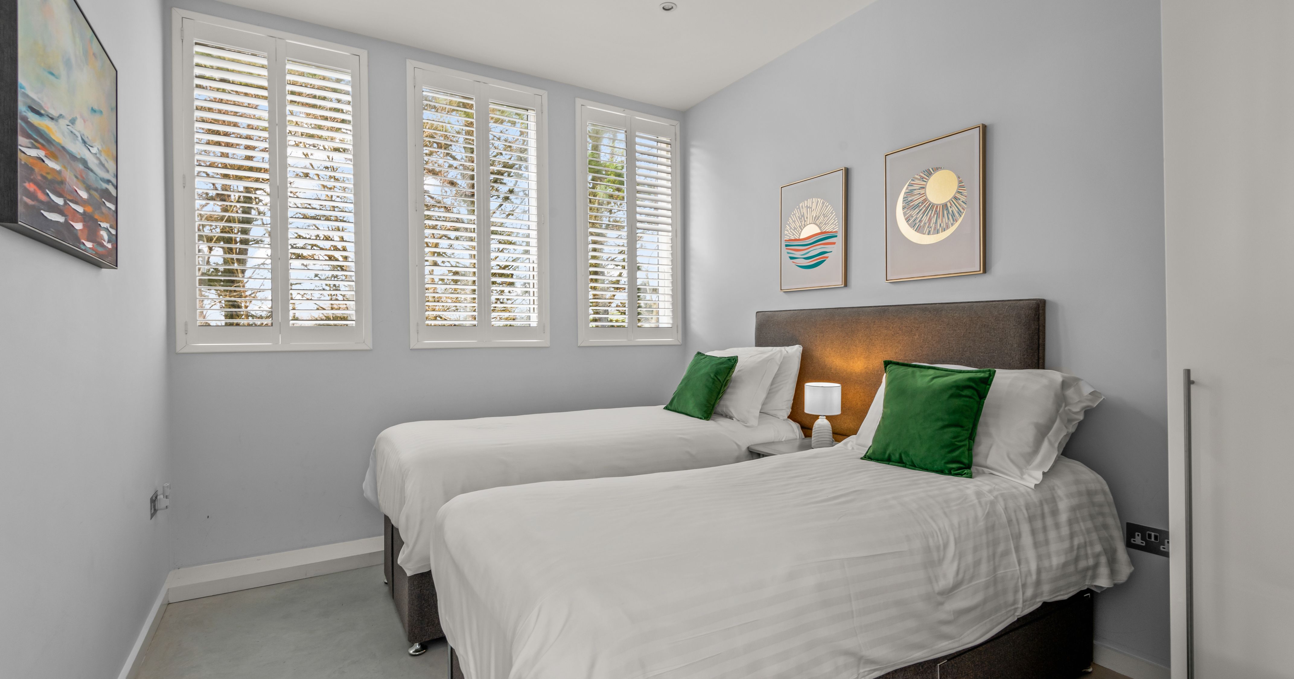 Modern bedroom with two single beds, green cushions, and large windows with white shutters