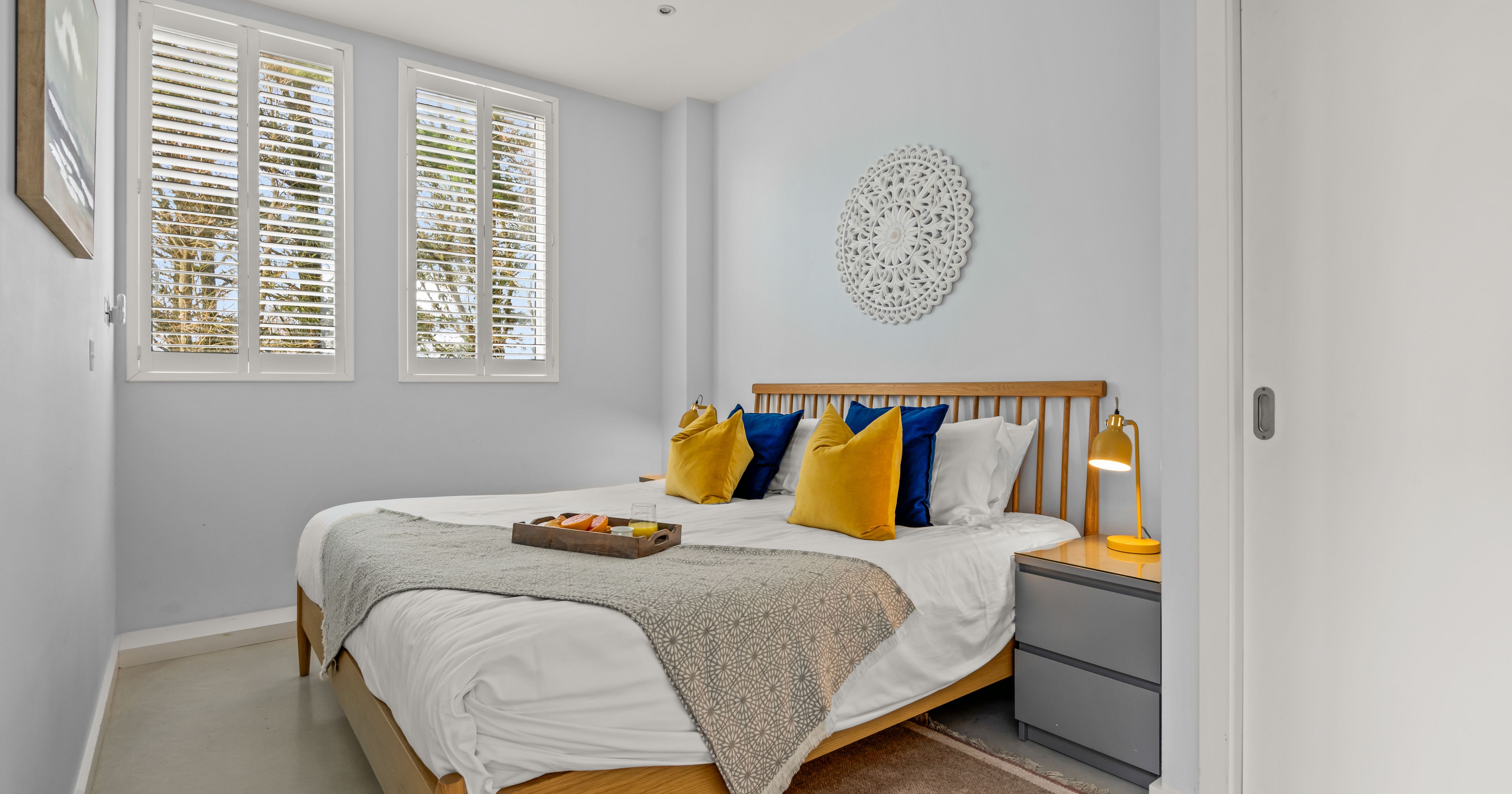 Modern bedroom with a wooden bed, blue and yellow pillows, large windows with shutters, and a nightstand with a yellow lamp.