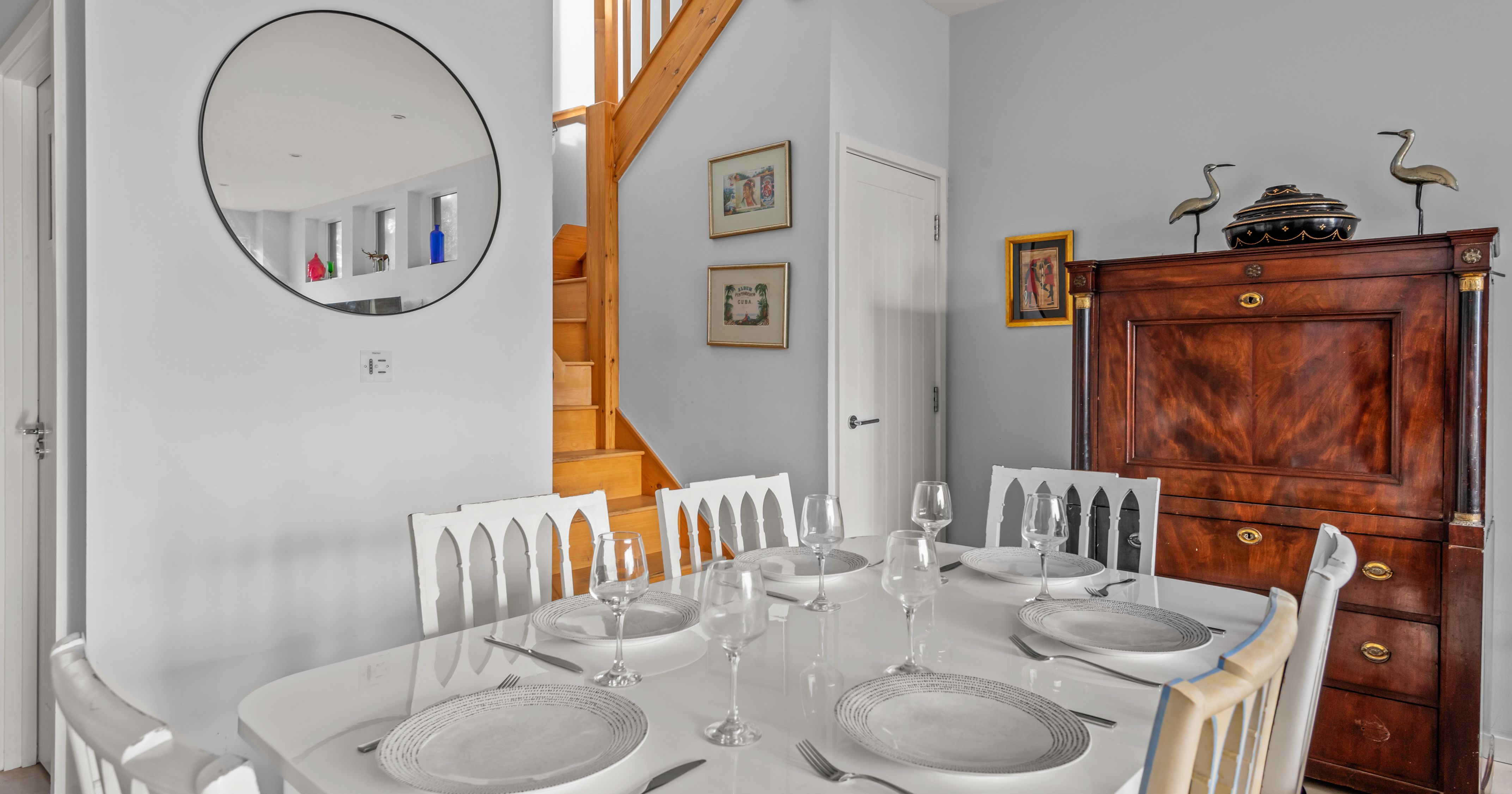 Elegant dining room with white table set for six, wooden staircase, and antique wooden furniture.